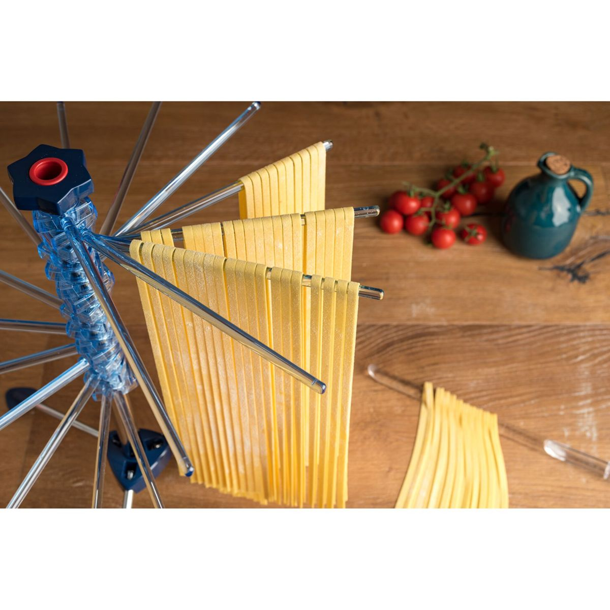 Fettuccine gialle fresche appese a un essiccatore blu, con pomodorini e una bottiglia verde.