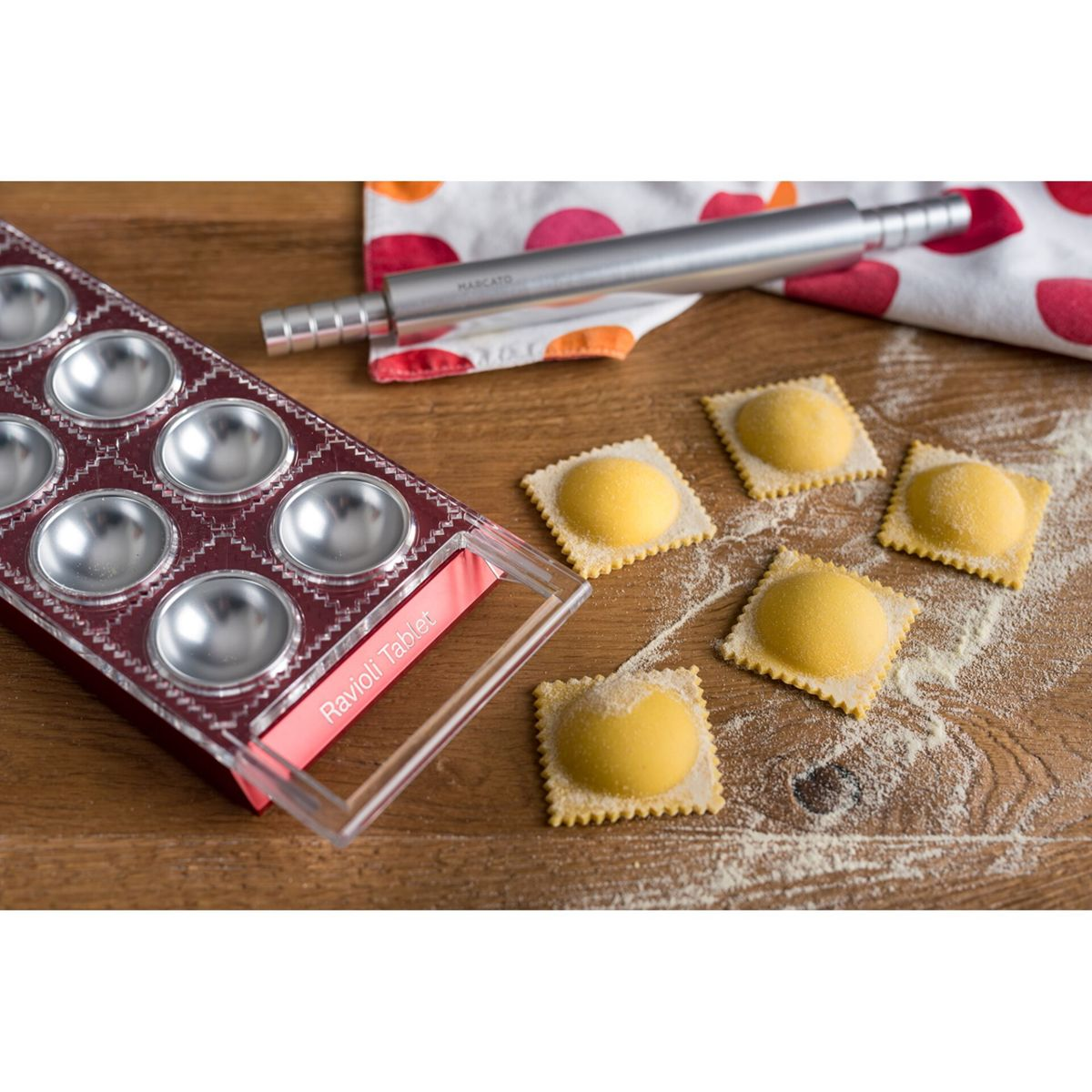 Eine rote Ravioli-Form, ein Nudelholz und frisch zubereitete, mit Mehl bestreute Ravioli auf einem Holztisch.