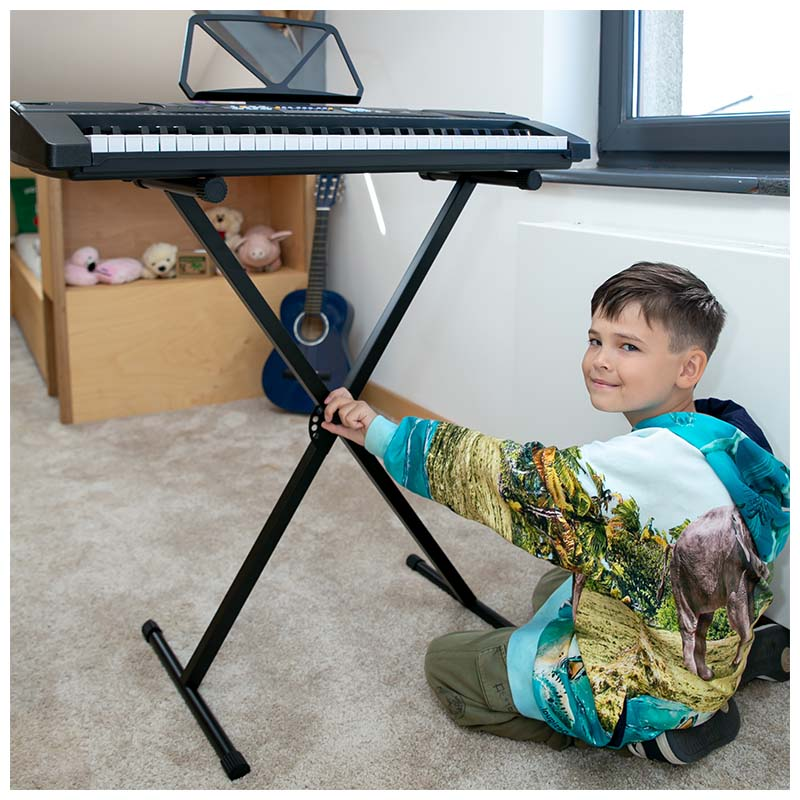 Ein Junge im bunten Hoodie kniet neben einem Keyboardständer auf einem Teppich in einem hellen Zimmer.
