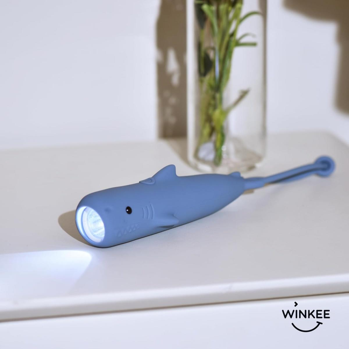 Eine blaue haiförmige Taschenlampe liegt auf einer weißen Oberfläche, das Licht brennt, im Hintergrund eine Vase mit Grün.