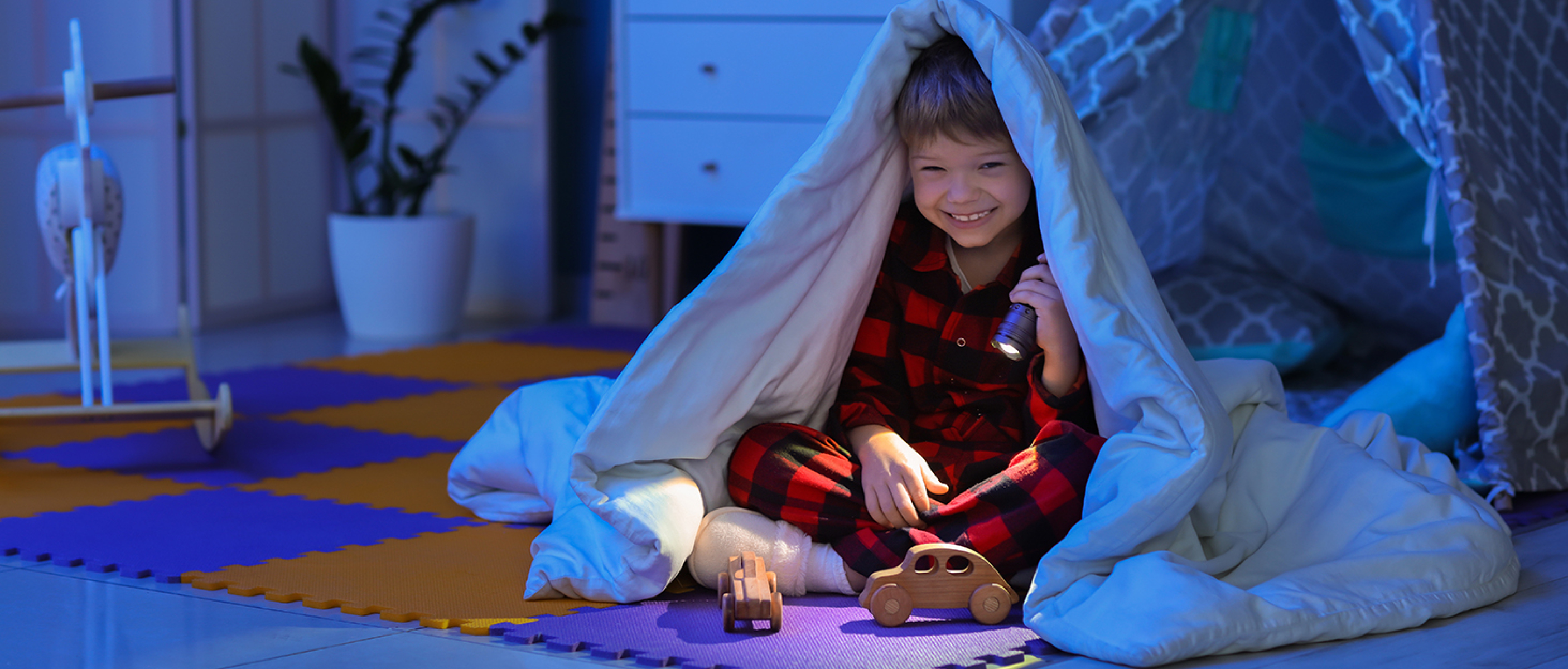 Ein lächelnder Junge in rot-schwarzem Pyjama sitzt mit einer Taschenlampe und Holzautos unter einer Decke.