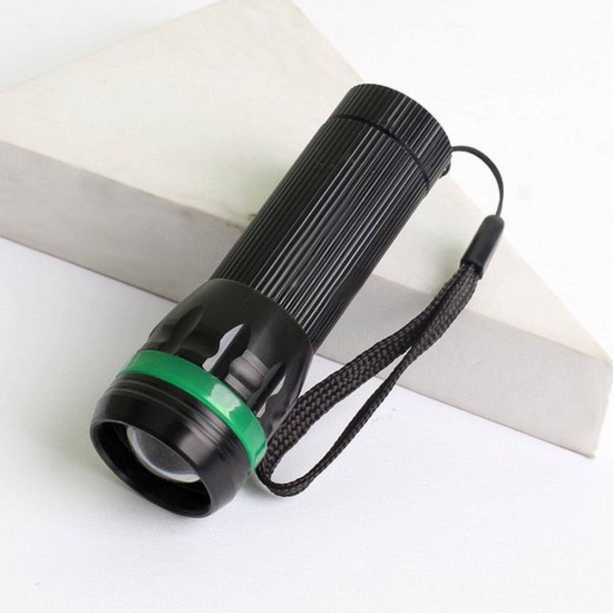 Eine schwarze Taschenlampe mit grünem Ring und Handschlaufe liegt auf einer weißen Oberfläche.