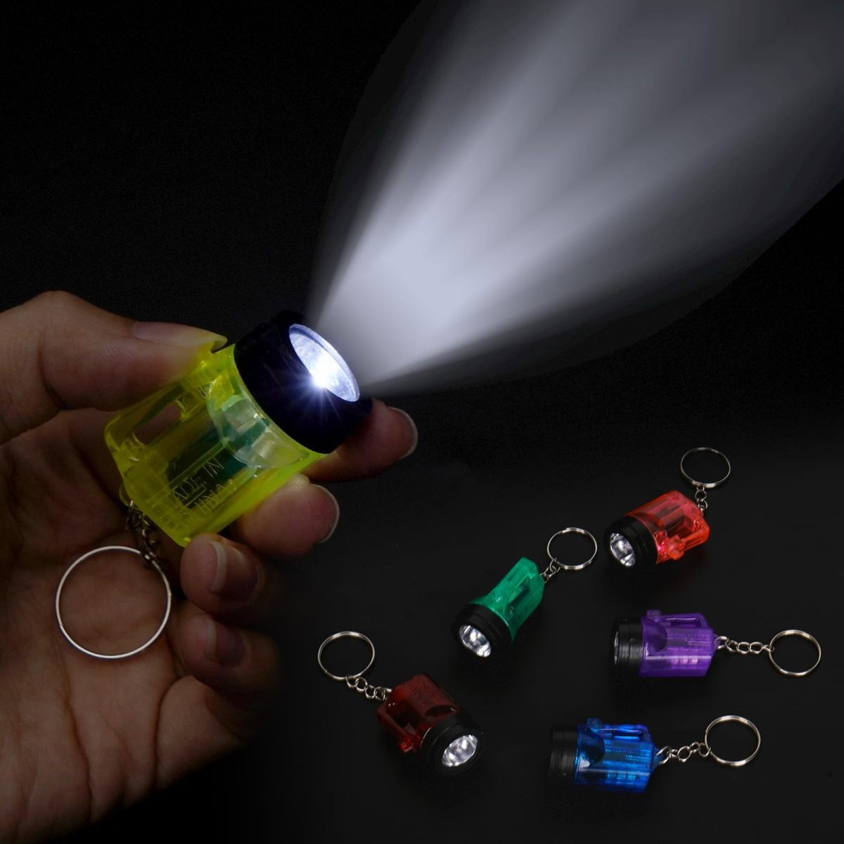 Eine Hand hält eine leuchtende gelbe Mini-Taschenlampe, mit mehreren anderen bunten Taschenlampen-Schlüsselanhängern.