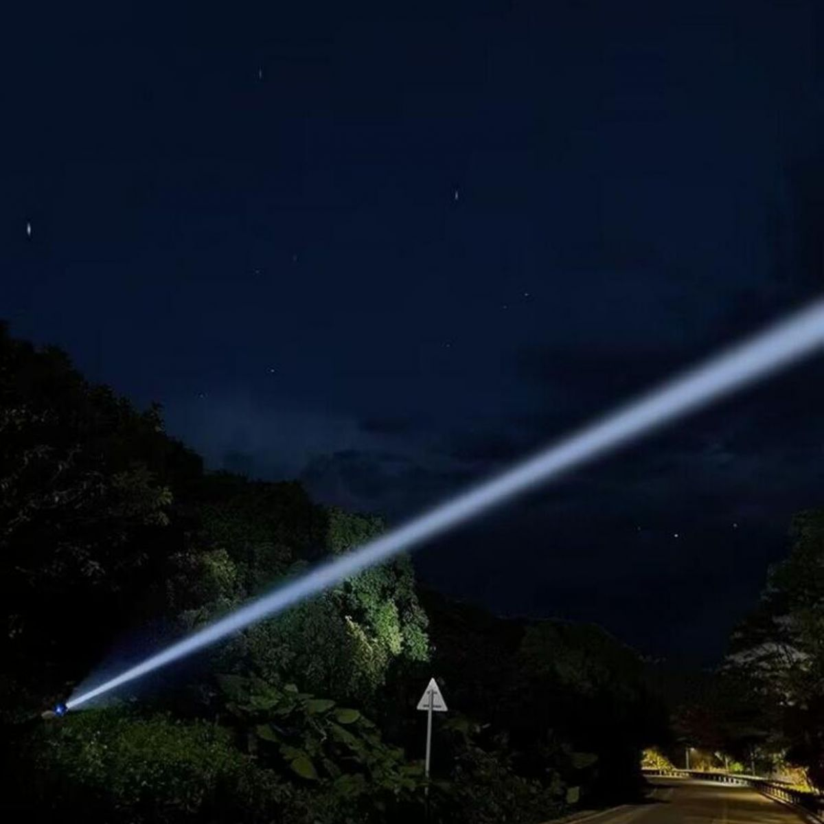 Ein starker Lichtkegel strahlt nachts von einer bewaldeten Straße in den dunklen Himmel.