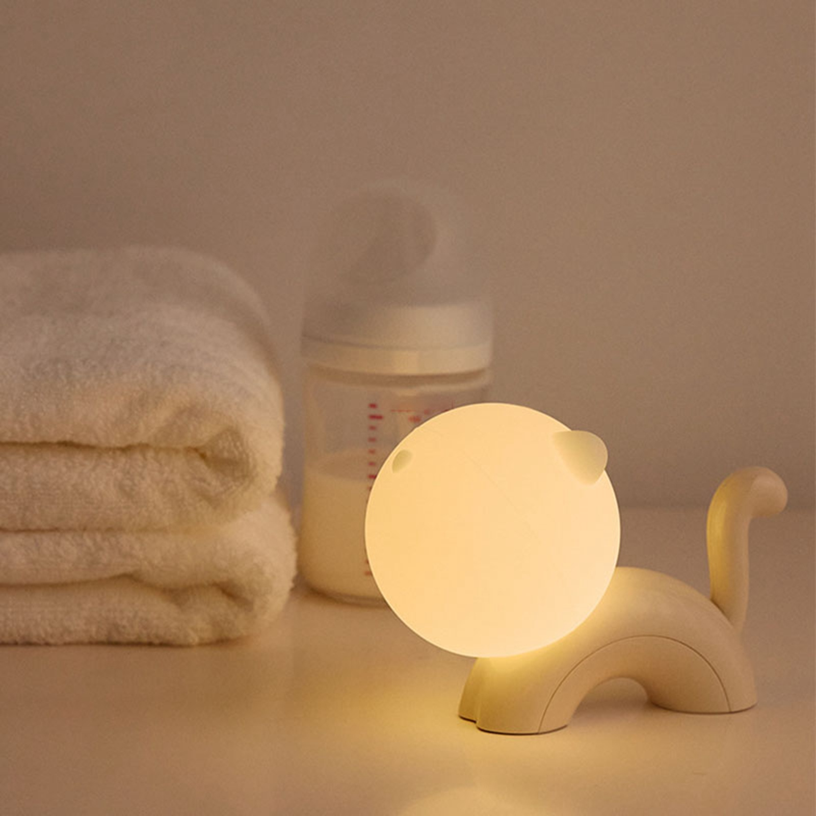 Eine katzenförmige Nachtlampe leuchtet auf einem Tisch, daneben eine Babyflasche und gefaltete Handtücher.