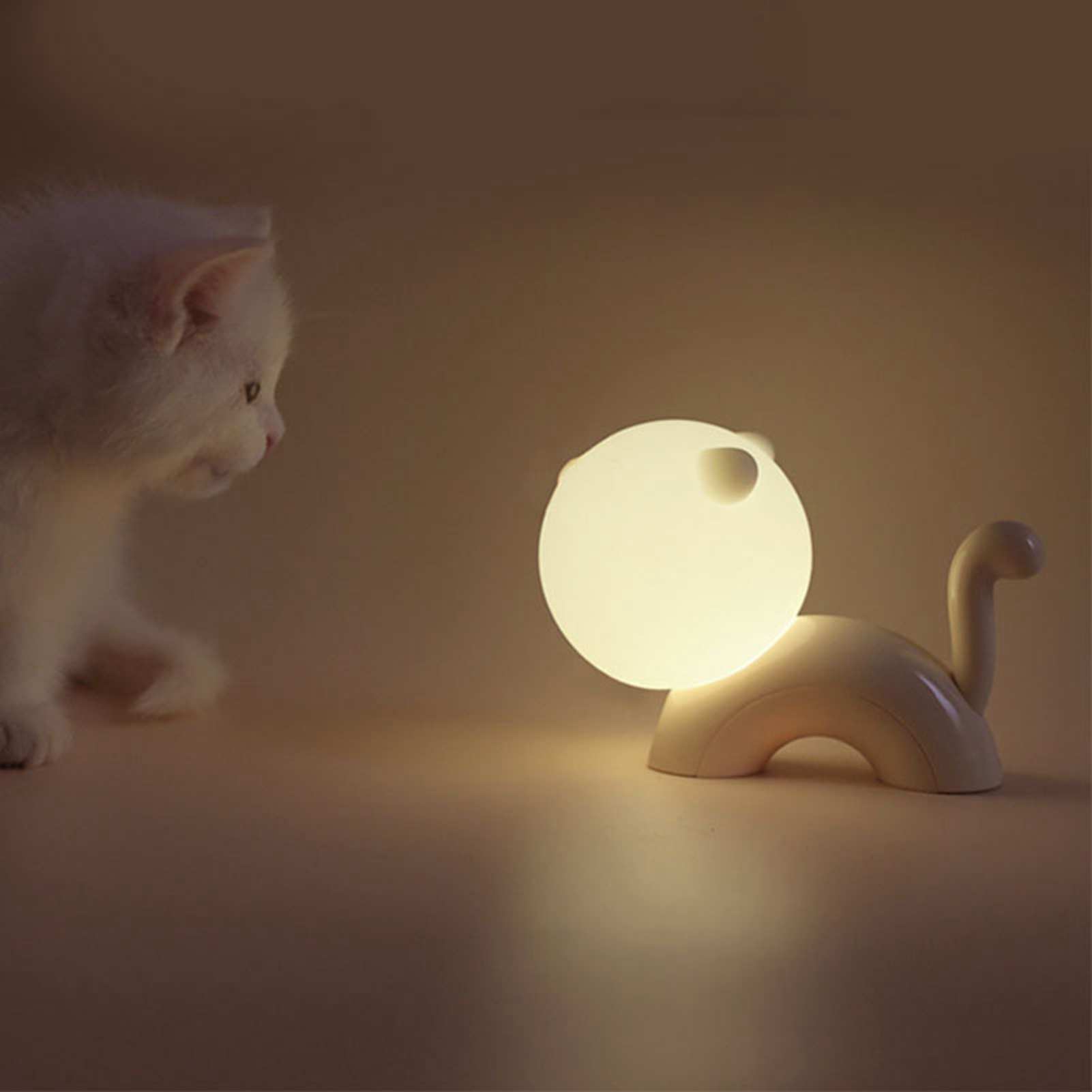 Ein weißes Kätzchen schaut neugierig auf eine katzenförmige Nachtlampe, die ein warmes Licht abgibt.