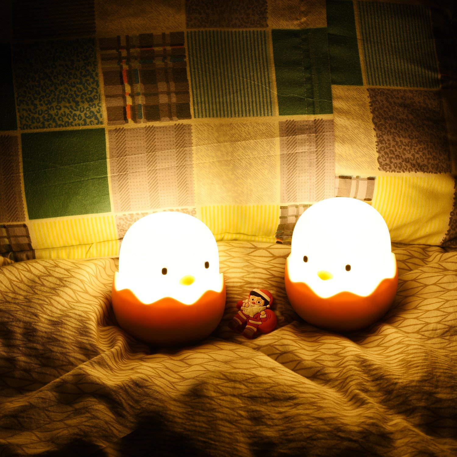 Zwei leuchtende Kükenlampen in Eierschalen stehen mit einer kleinen Weihnachtsmannfigur auf einem Bett vor einem Kissen.