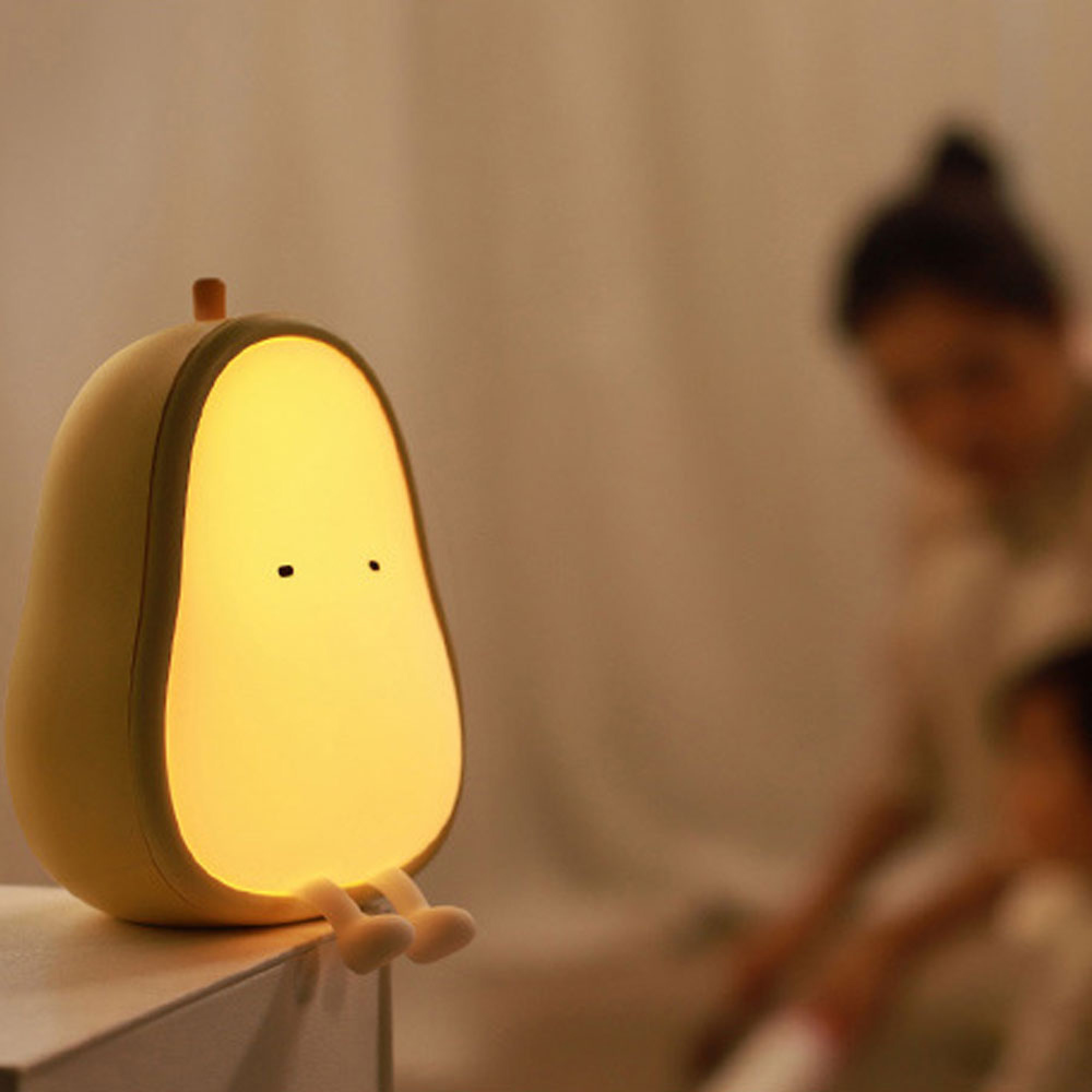Eine Birnenförmige, leuchtende Lampe mit kleinen Beinen steht auf einem Tisch, eine Person sitzt unscharf im Hintergrund.