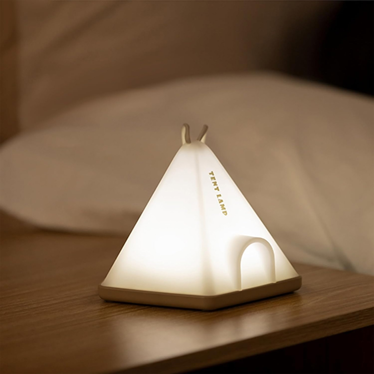 Eine pyramidenförmige Zeltlampe leuchtet warm auf einem Holz-Nachtkästchen mit einem Kissen im Hintergrund.