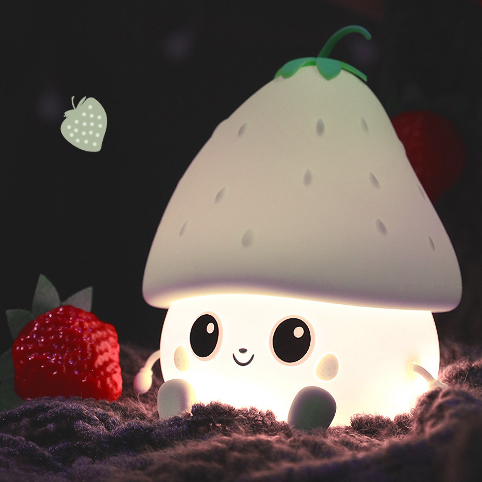 Eine süße, leuchtende erdbeerförmige Lampe mit Gesicht sitzt auf einer Decke neben echten Erdbeeren.