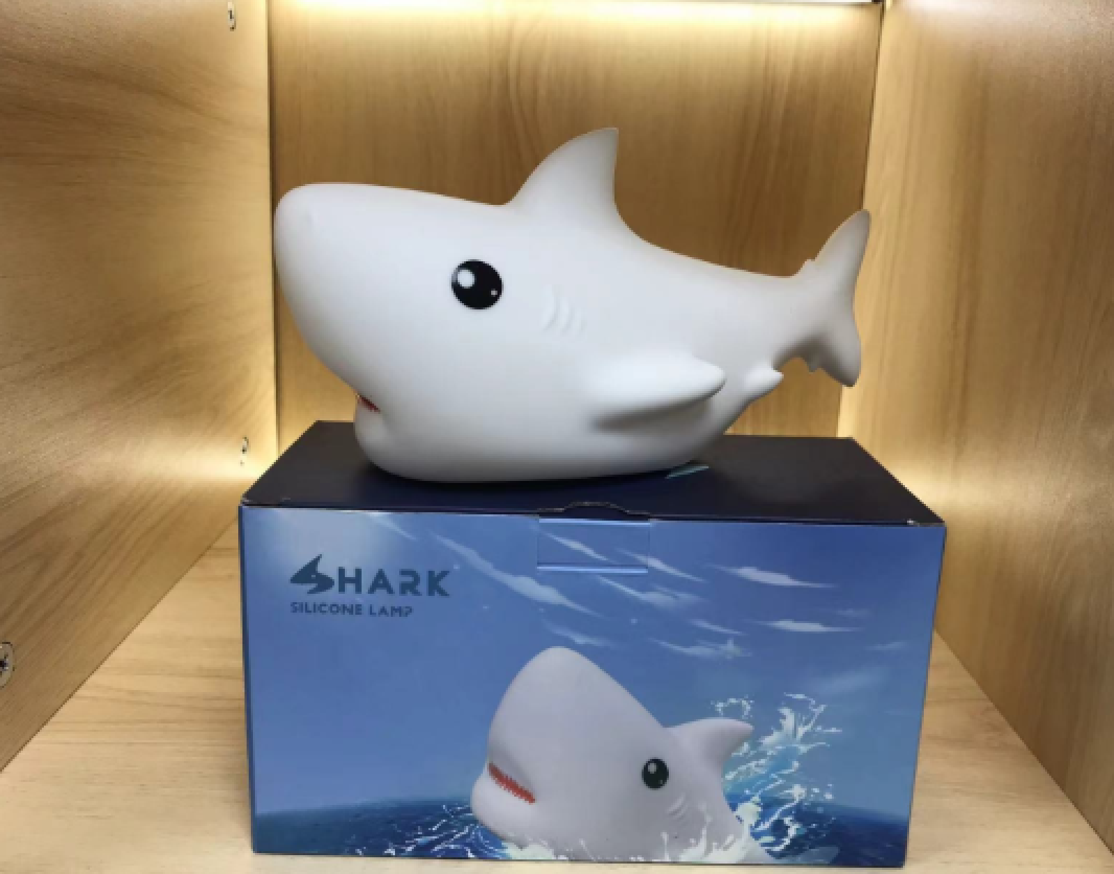 Eine weiße Haifisch-Silikonlampe sitzt auf einer blauen Verpackungsbox mit Haifisch-Design in einem Holzregal.