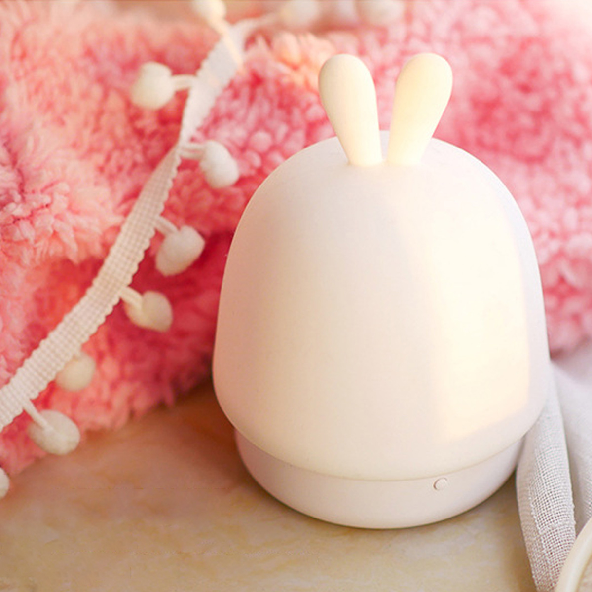 Eine kleine, weiße Hasenlampe steht vor einer rosa Decke mit Pomponborte.