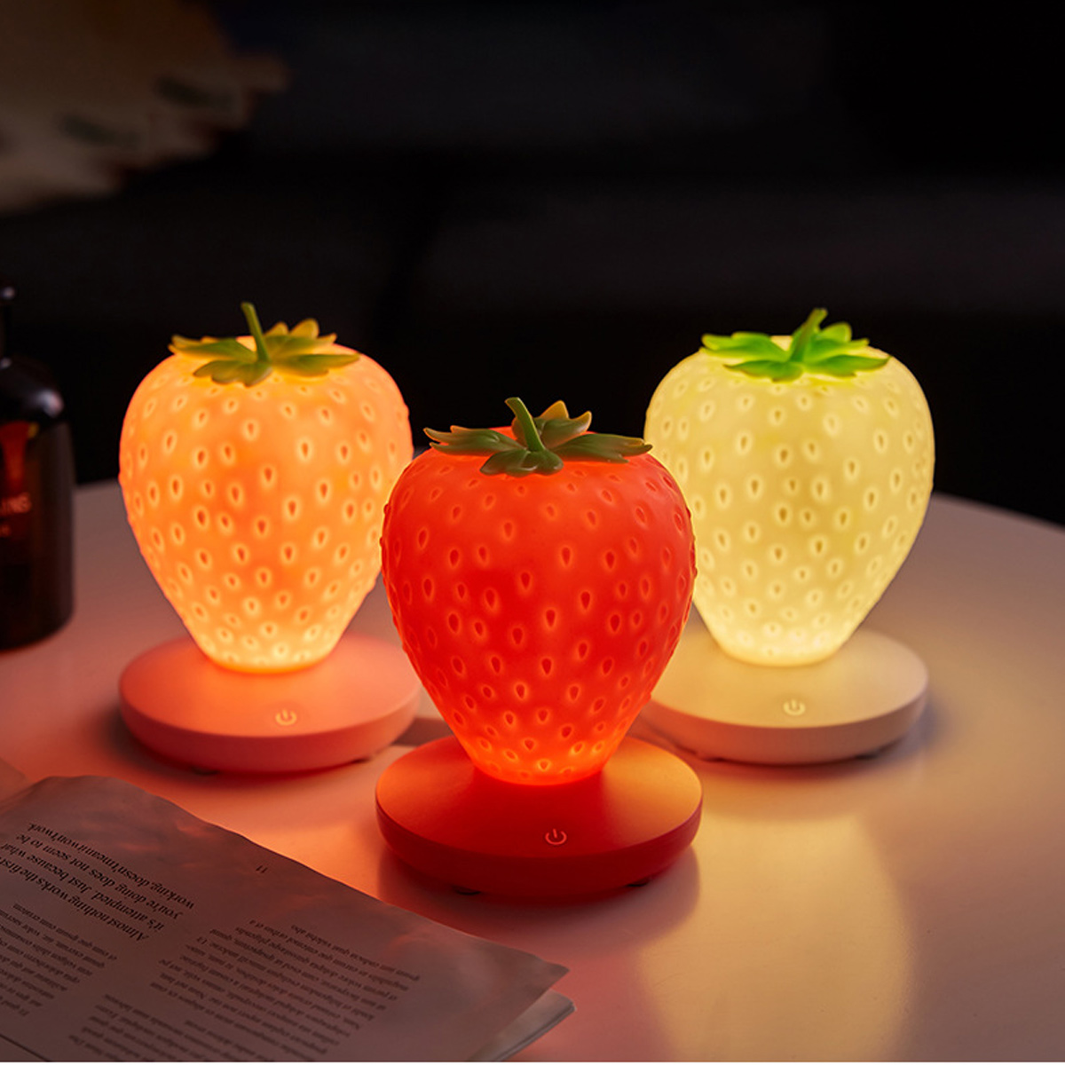 Drei erdbeerförmige Nachtlichter leuchten in sanften Farben auf einem weißen Tisch.