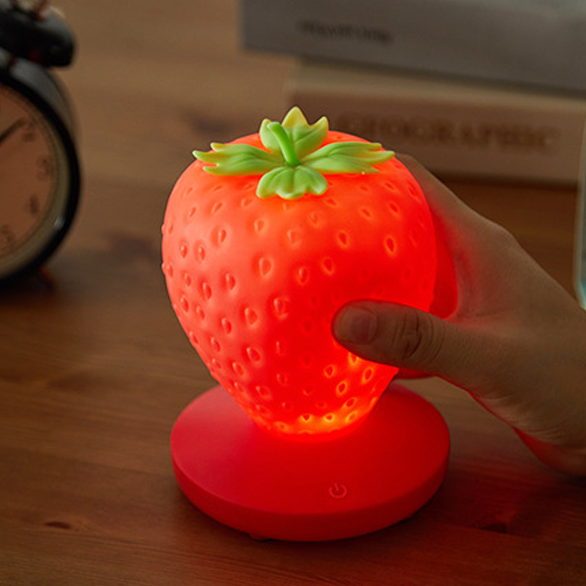 Eine Hand berührt eine leuchtende, erdbeerförmige Lampe auf einem Holztisch mit Uhr und Büchern im Hintergrund.