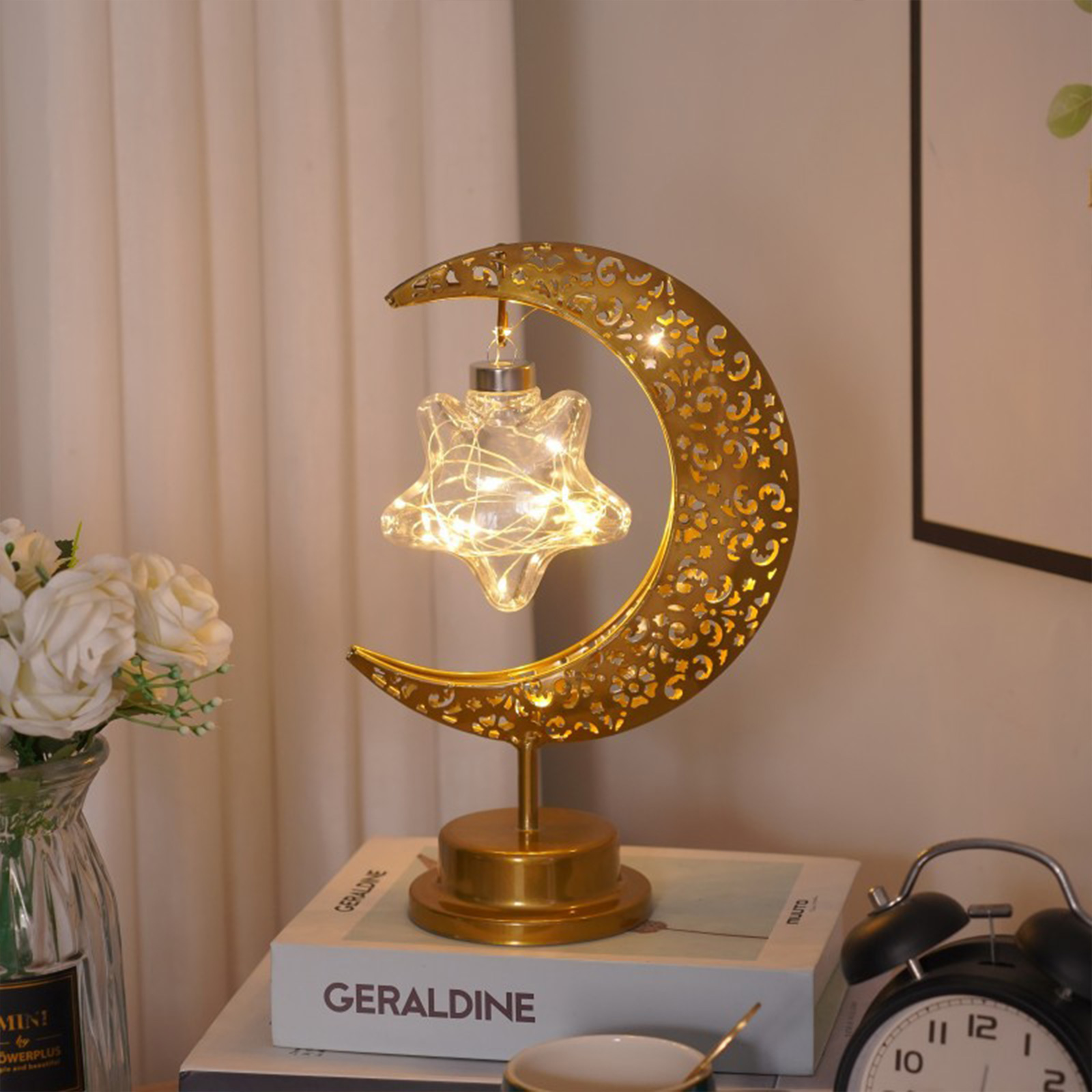 Eine dekorative goldene Mondsichel-Lampe mit beleuchtetem Stern auf einem Buch neben Blumen und einer Uhr.