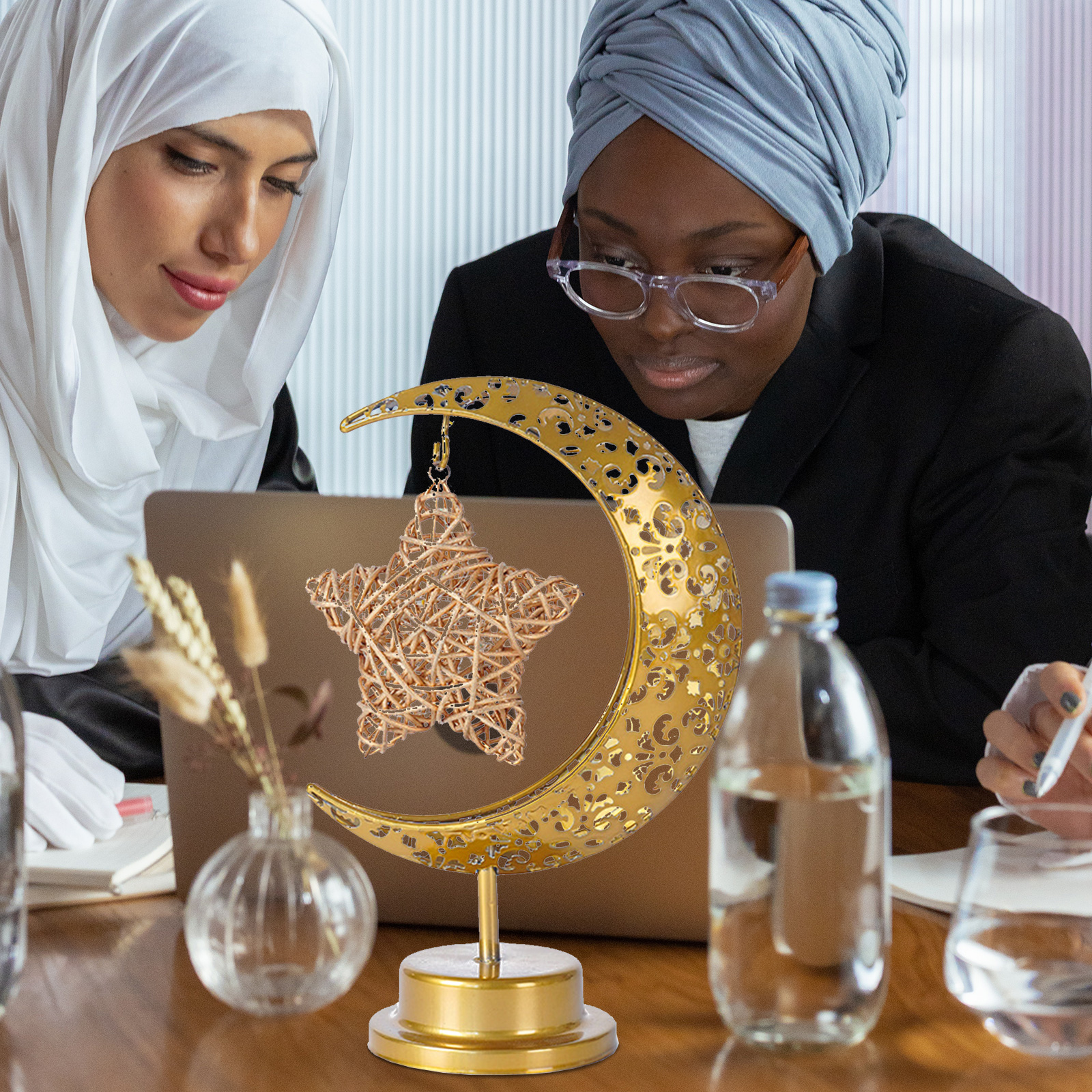Zwei Frauen im Büro schauen auf ein Laptop, davor eine goldene Mondsichel mit Stern und eine Wasserflasche.