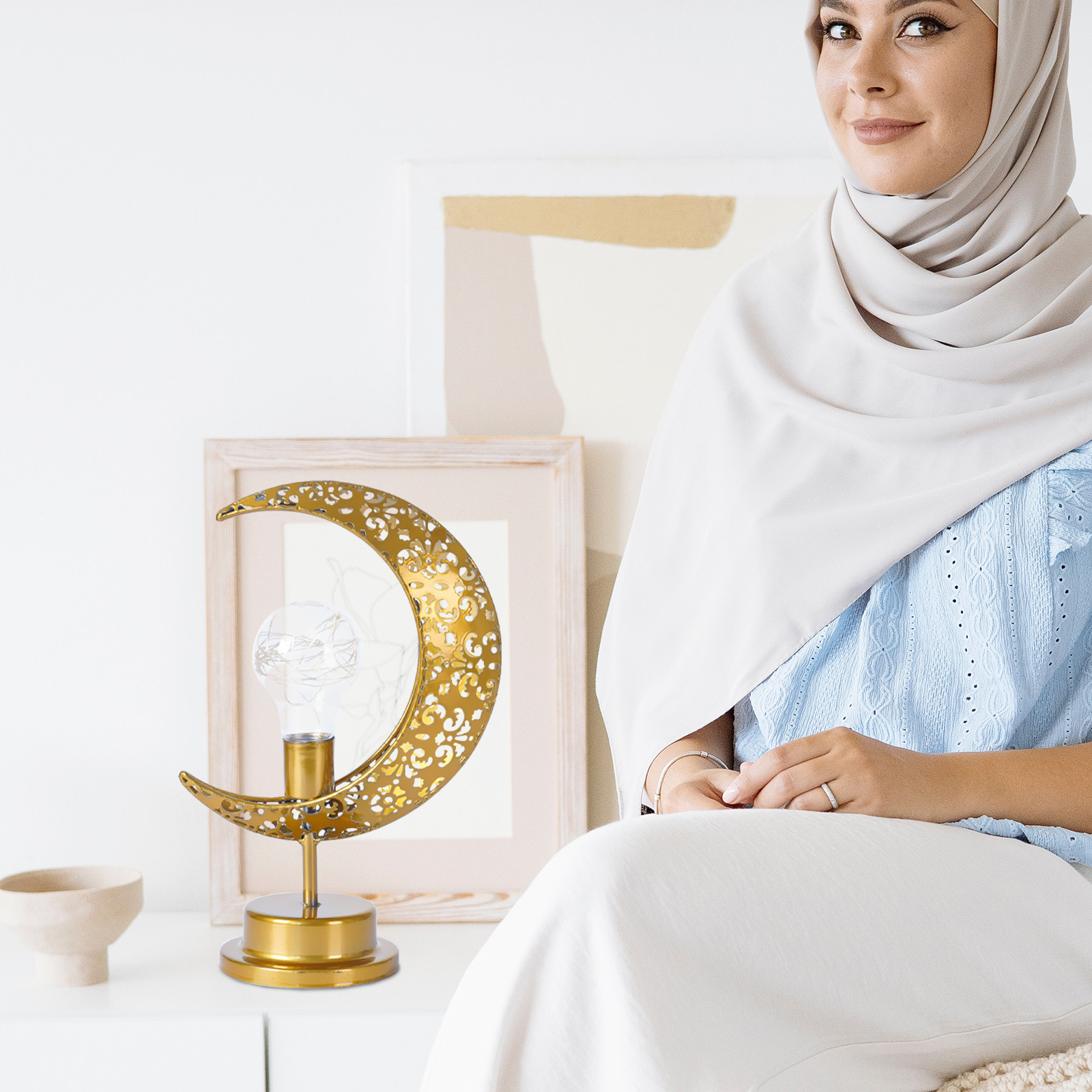 Eine Frau in weißem Hijab lächelt neben einer goldenen mondförmigen Tischlampe auf einem weißen Tisch.