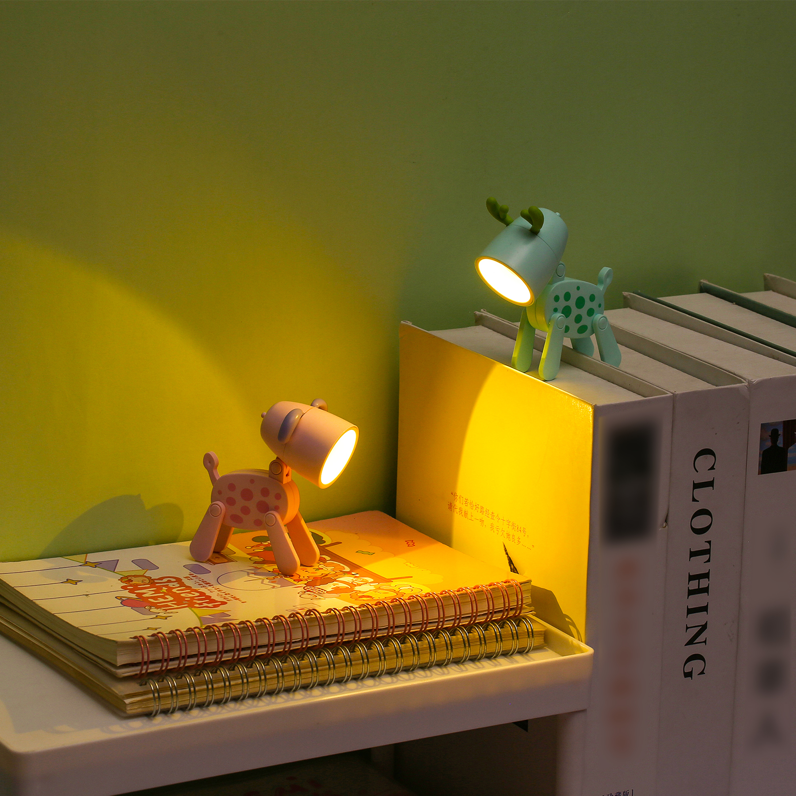 Zwei kleine tierförmige LED-Lampen leuchten warm auf gestapelten Büchern vor einer grünen Wand.