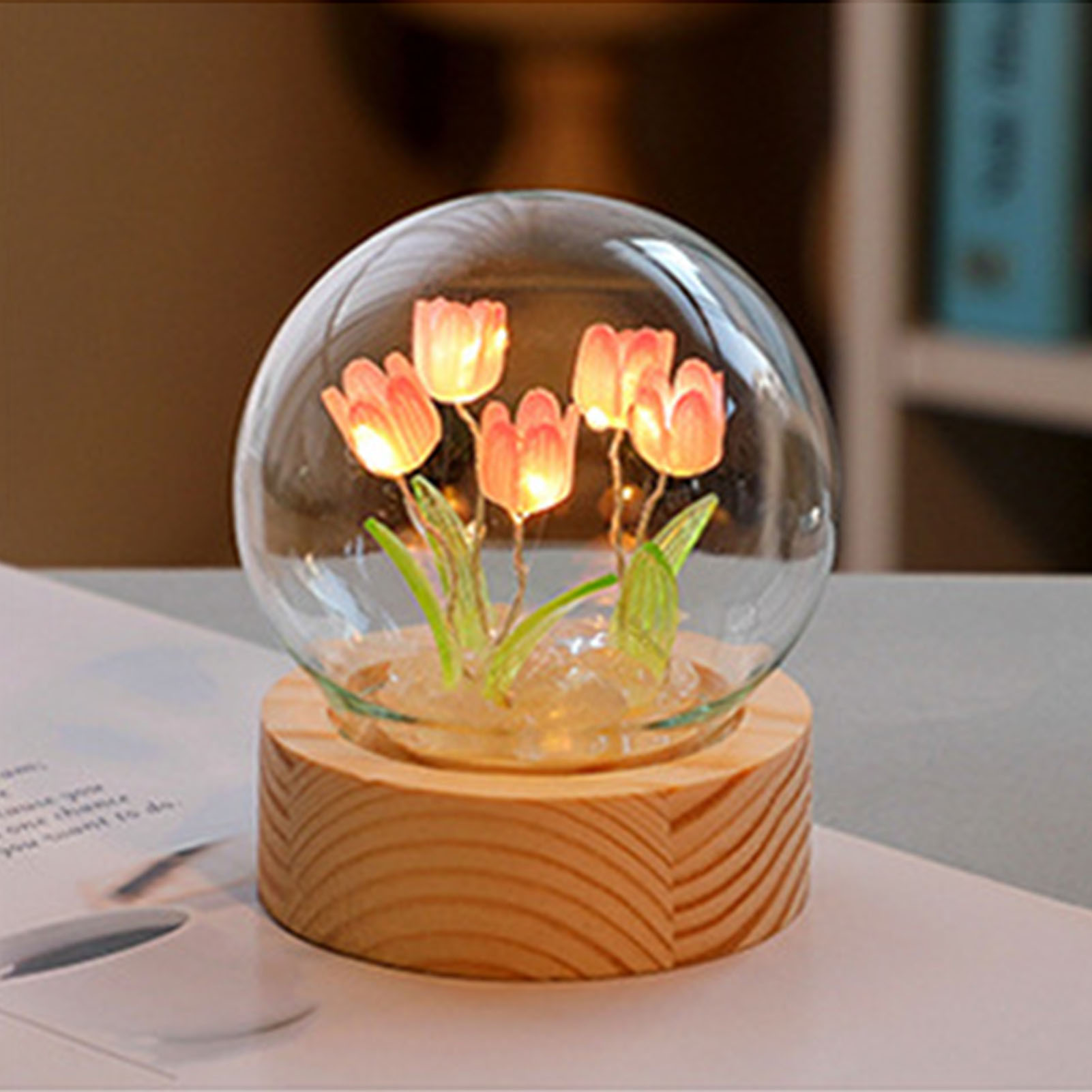 Eine Glaskugel mit beleuchteten rosafarbenen Tulpen auf einem Holzsockel auf einem Tisch.