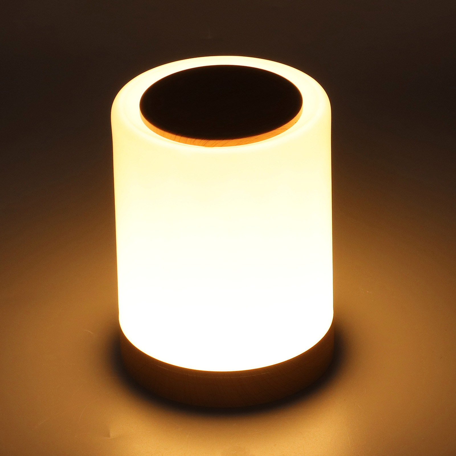 Eine zylindrische weiße Lampe mit Holzsockel und Oberseite strahlt warmes Licht vor dunklem Hintergrund.