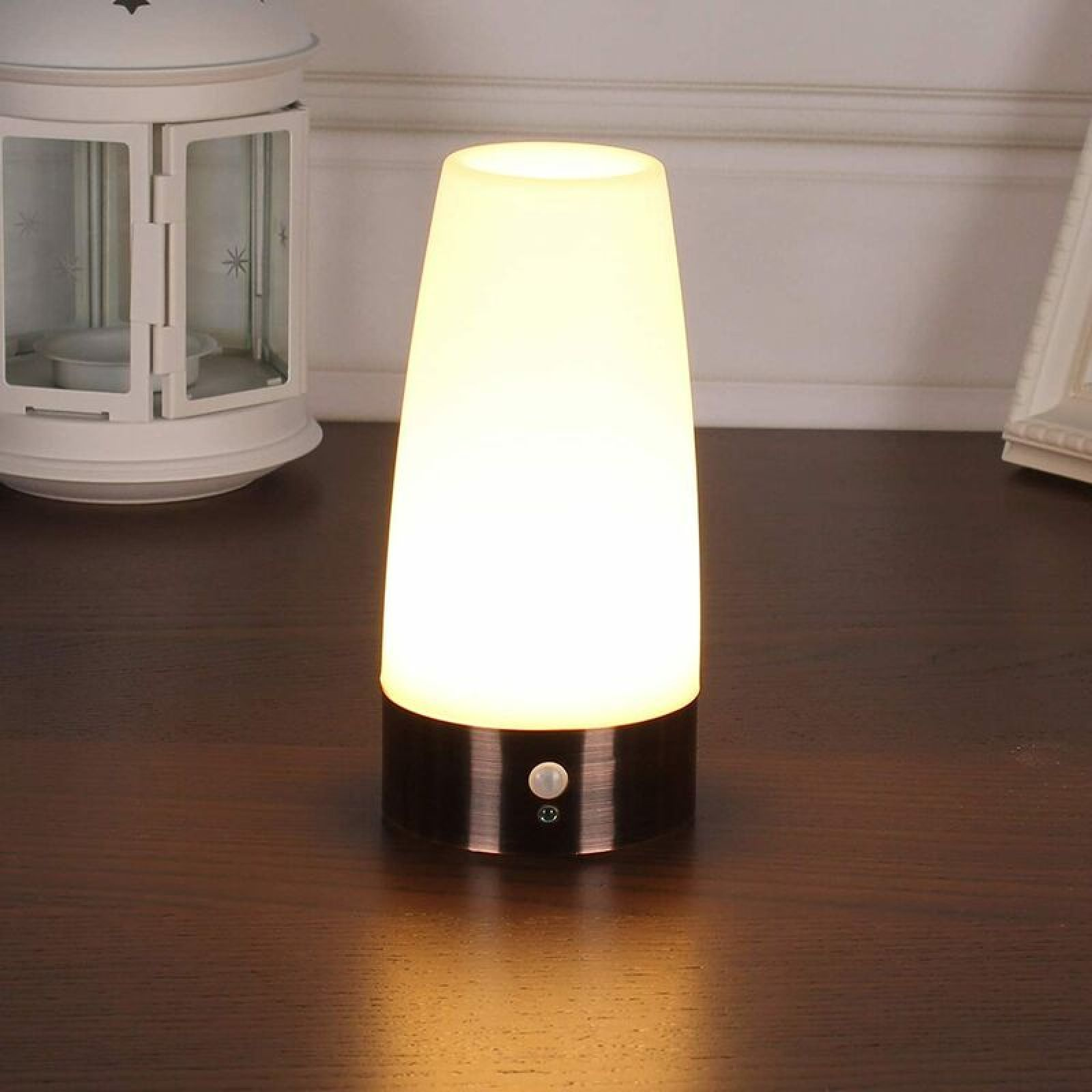 Eine weiß leuchtende Tischlampe mit schwarzem Sockel steht auf einem braunen Holztisch vor einer hellen Wand.