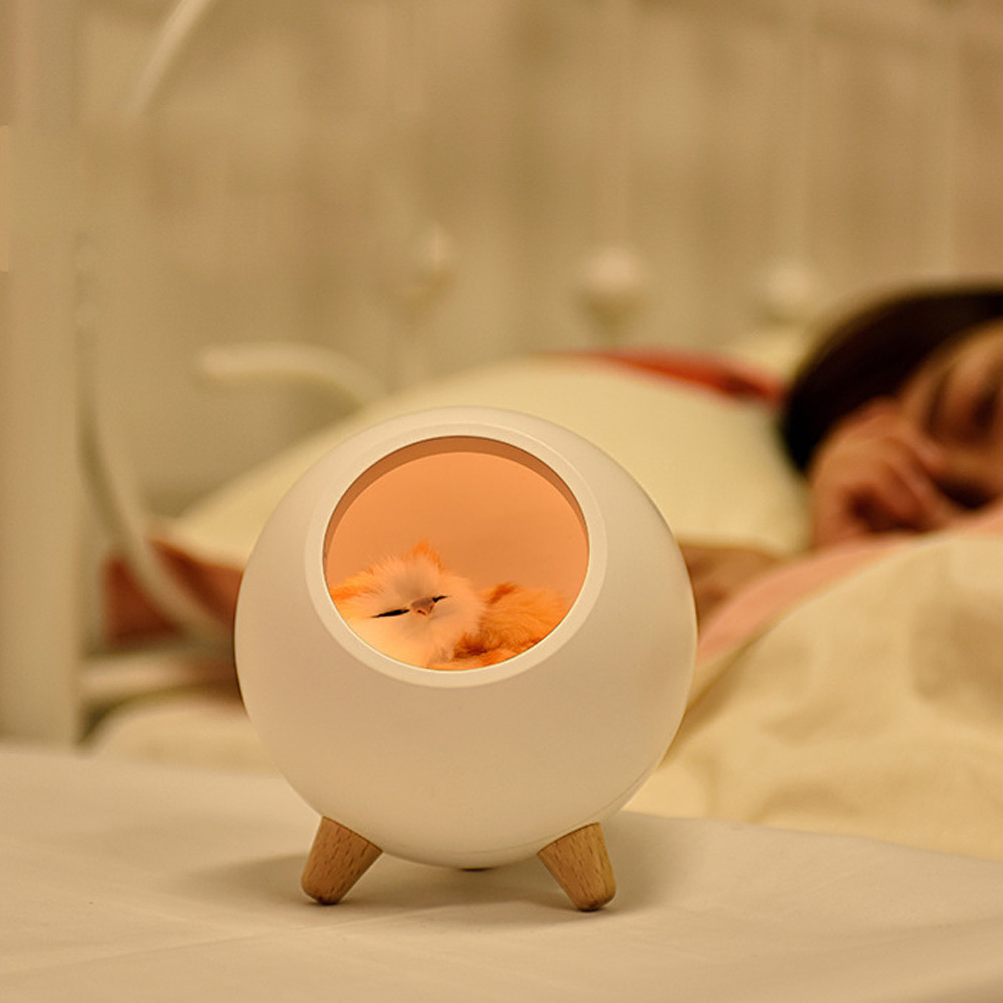 Eine kleine Lampe auf drei Beinen mit einer schlafenden Katze im warmen Licht, im Hintergrund liegt jemand im Bett.