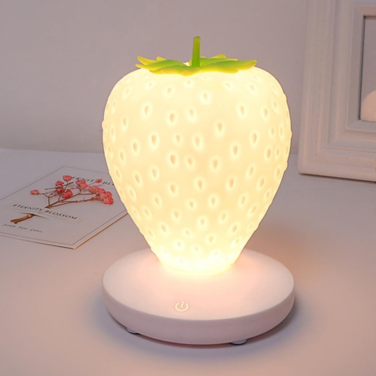 Eine leuchtende Erdbeeren-Lampe auf einem weißen Sockel, umgeben von einem kleinen Bild und einer Karte auf dem Tisch.