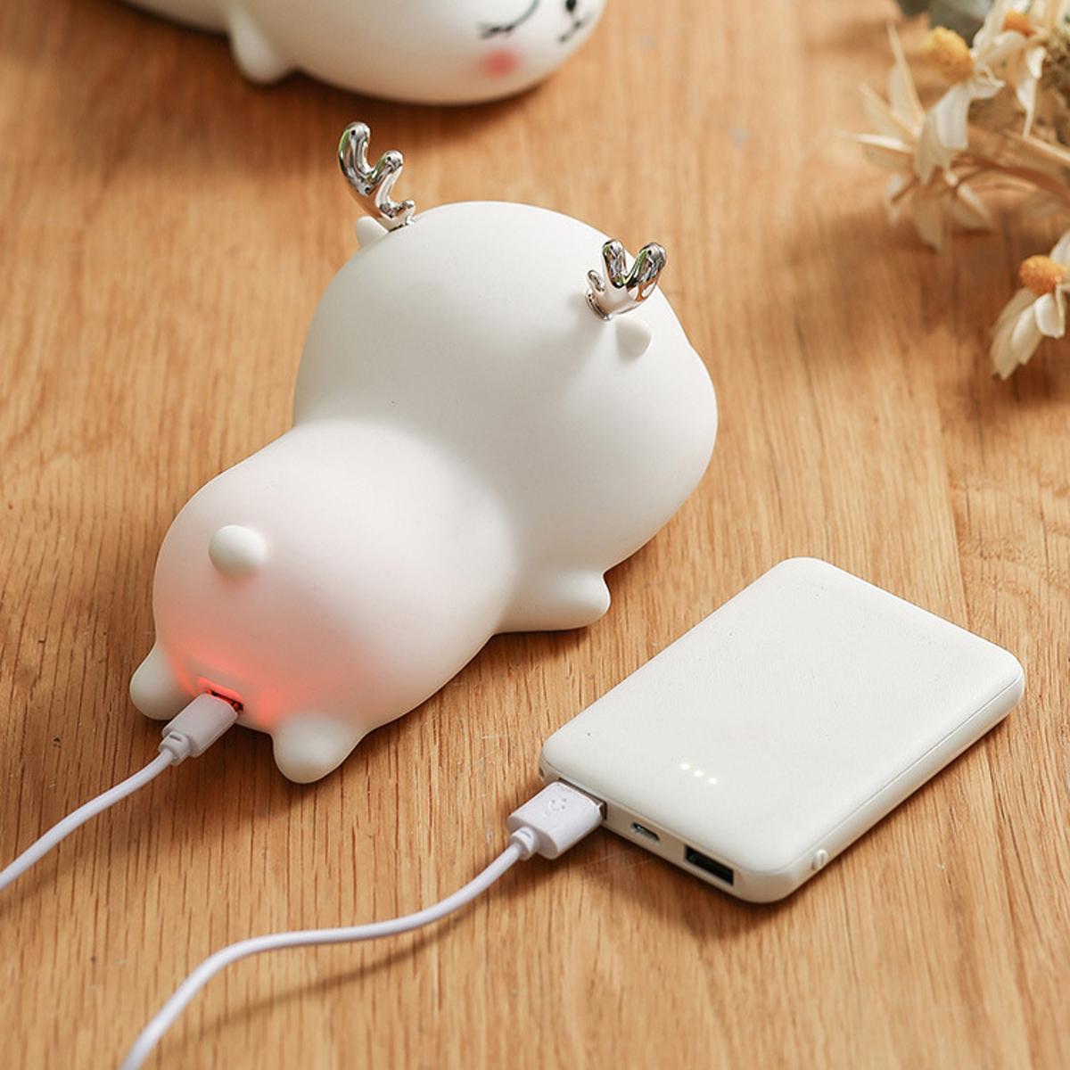 Ein weißes, tierähnliches Nachtlicht mit silbernen Geweihen wird von einer weißen Powerbank auf einem Holztablett aufgeladen.