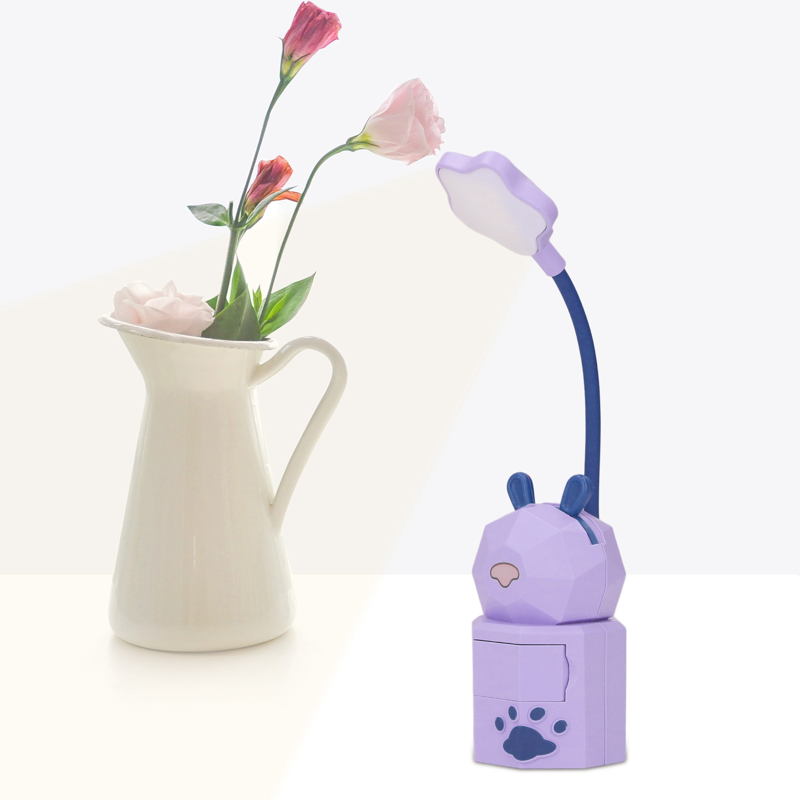 Ein weißer Krug mit rosa Blumen steht neben einer lila Schreibtischlampe in Tierform vor weißem Hintergrund.