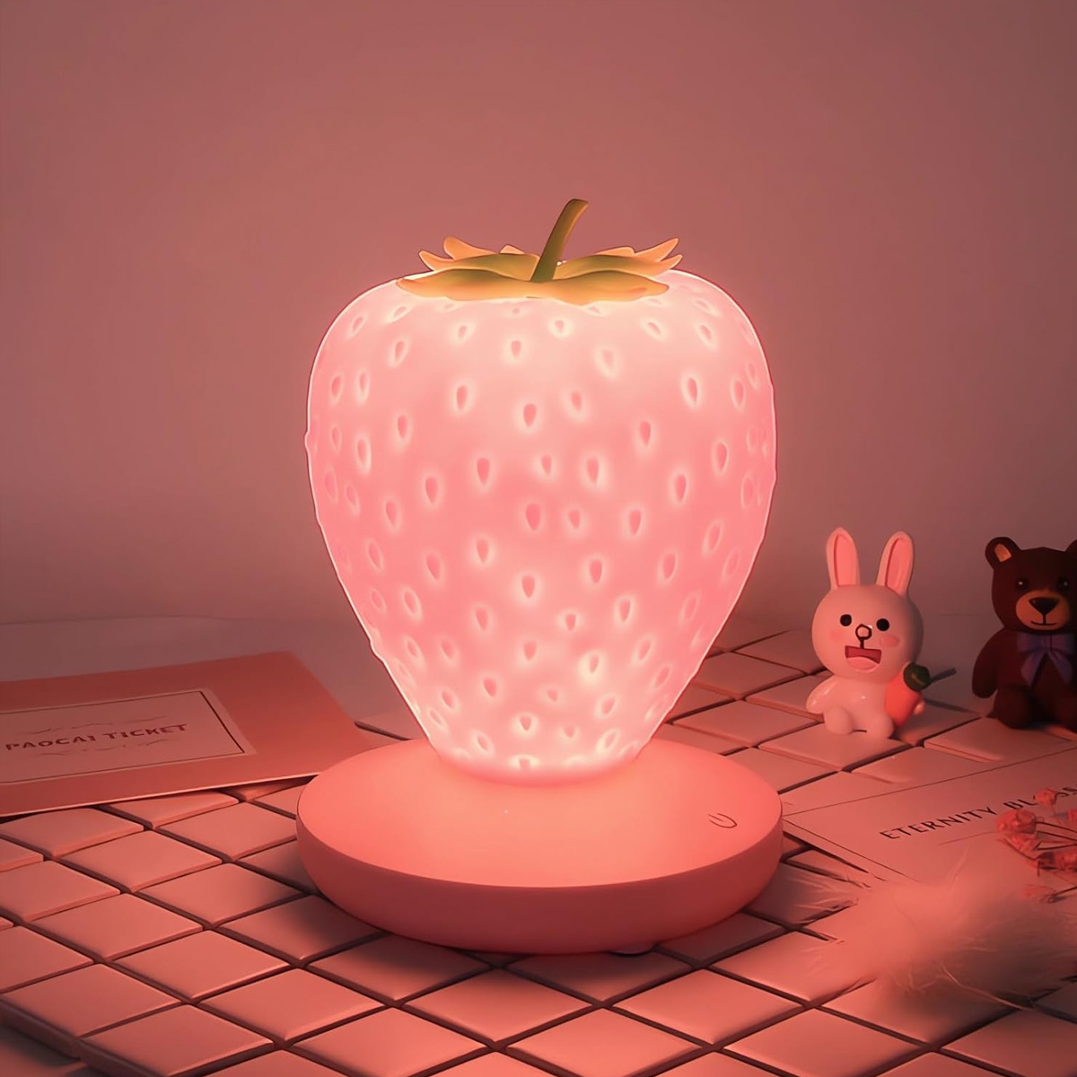 Eine pinke Erdbeertischlampe auf einem Tisch, flankiert von kleinen Spielzeugen vor einem schlichten Hintergrund.