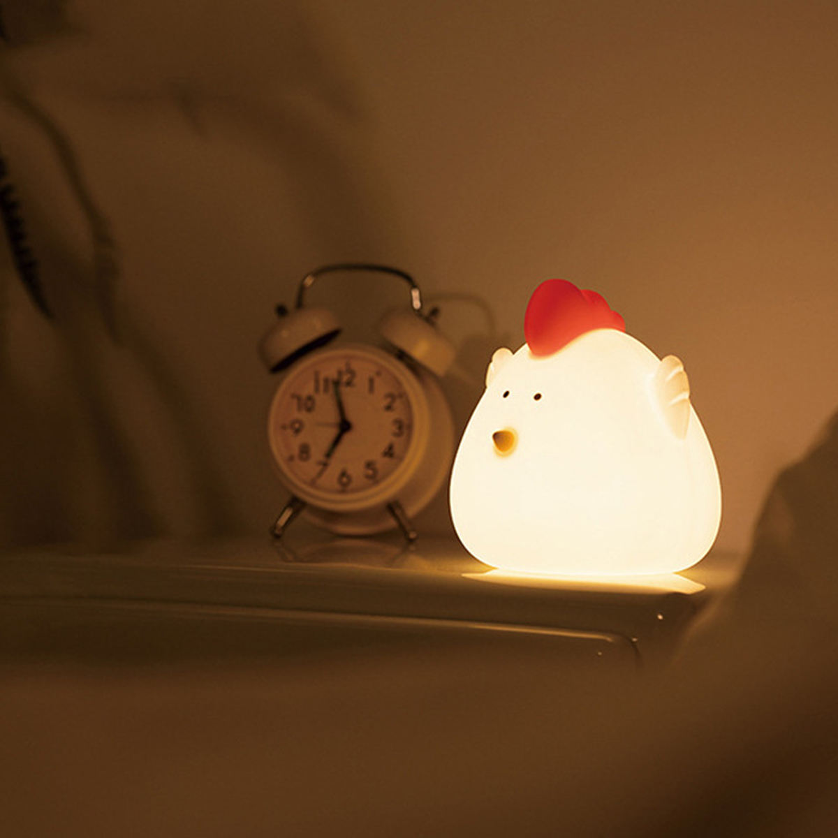 Eine leuchtende Hühner-Nachtlampe und ein weißer Wecker stehen auf einem Tisch im gedimmten Licht.
