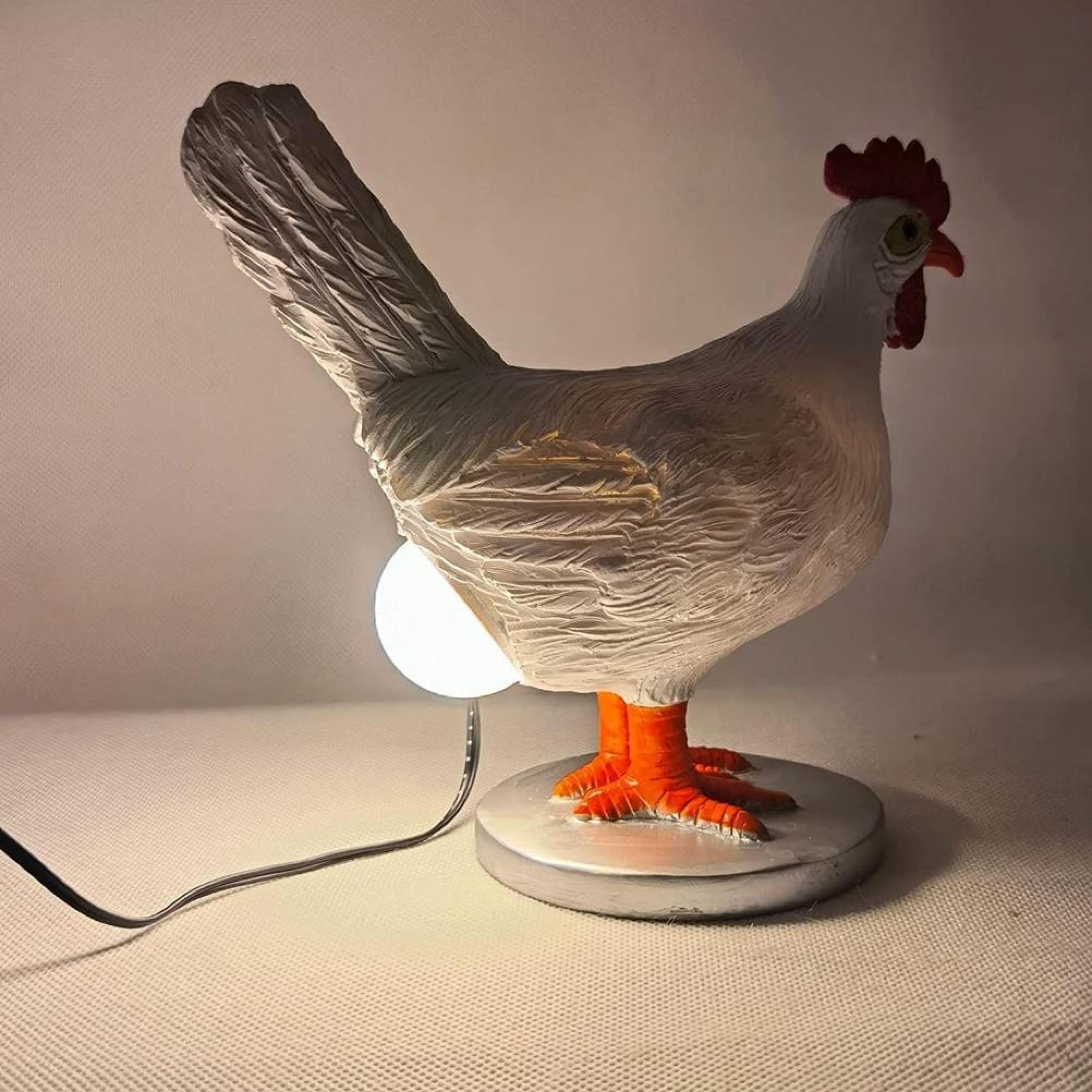 Eine weiße Hühnerlampe mit rotem Kamm und orangefarbenen Beinen steht auf einer runden Platte; eine Glühbirne leuchtet.