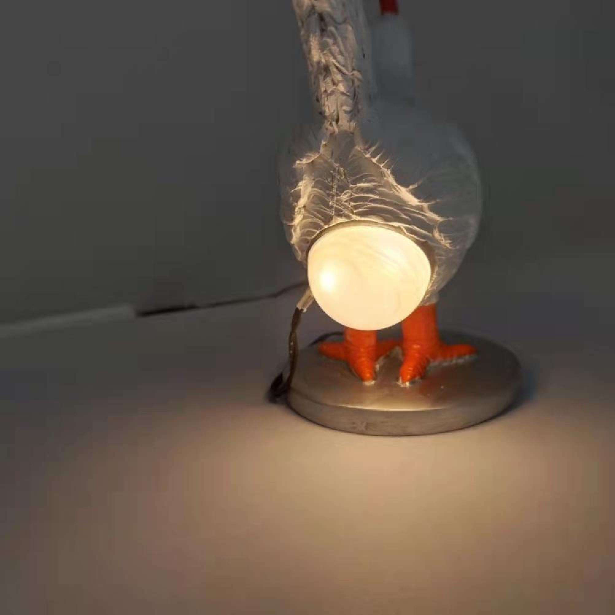 Eine skurrile weiße Hühnerlampe mit orangen Beinen steht auf einem runden Sockel und leuchtet hinten.