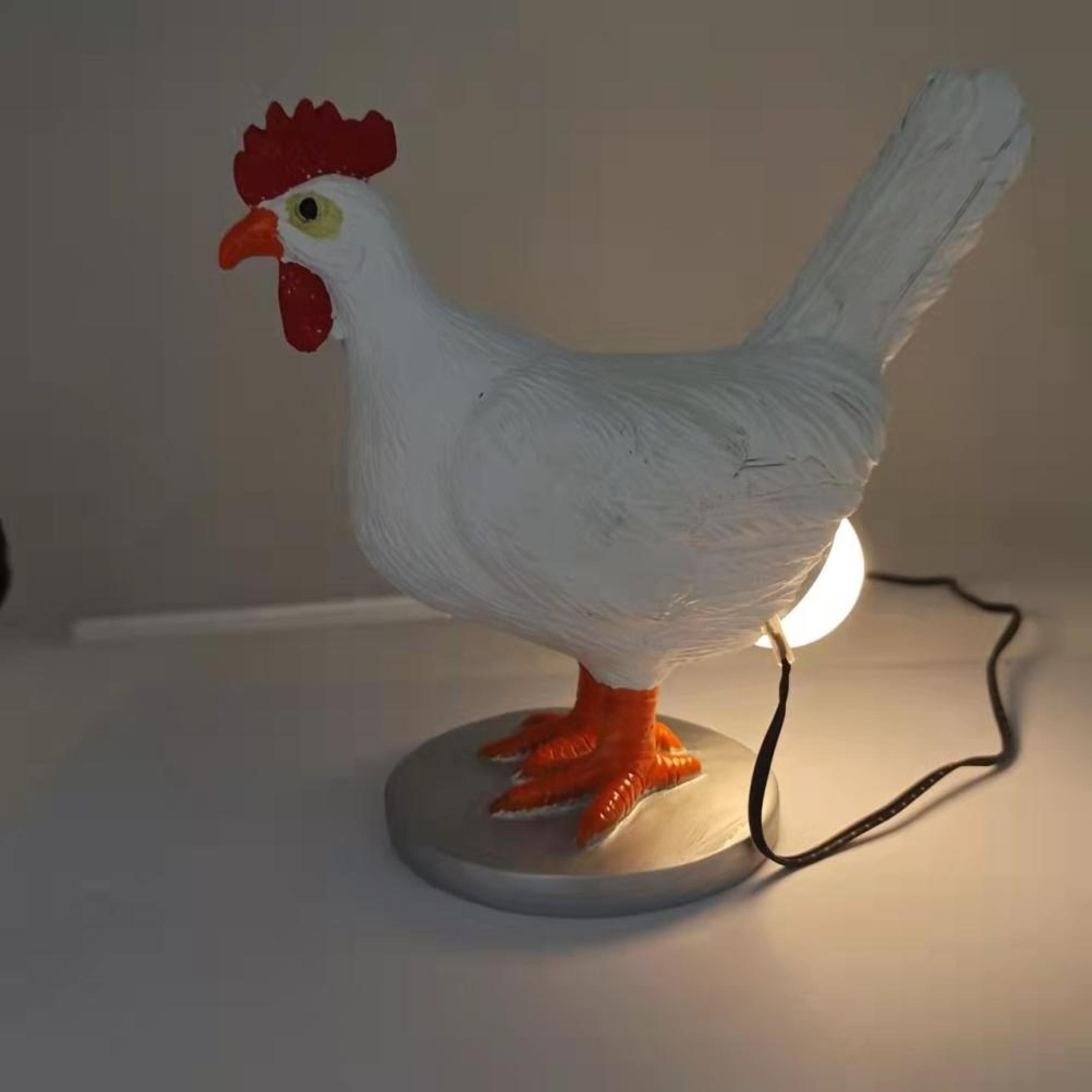 Eine weiße Hahnenlampe mit rotem Kamm und orangefarbenen Beinen steht auf einer grauen Basis und leuchtet von hinten.