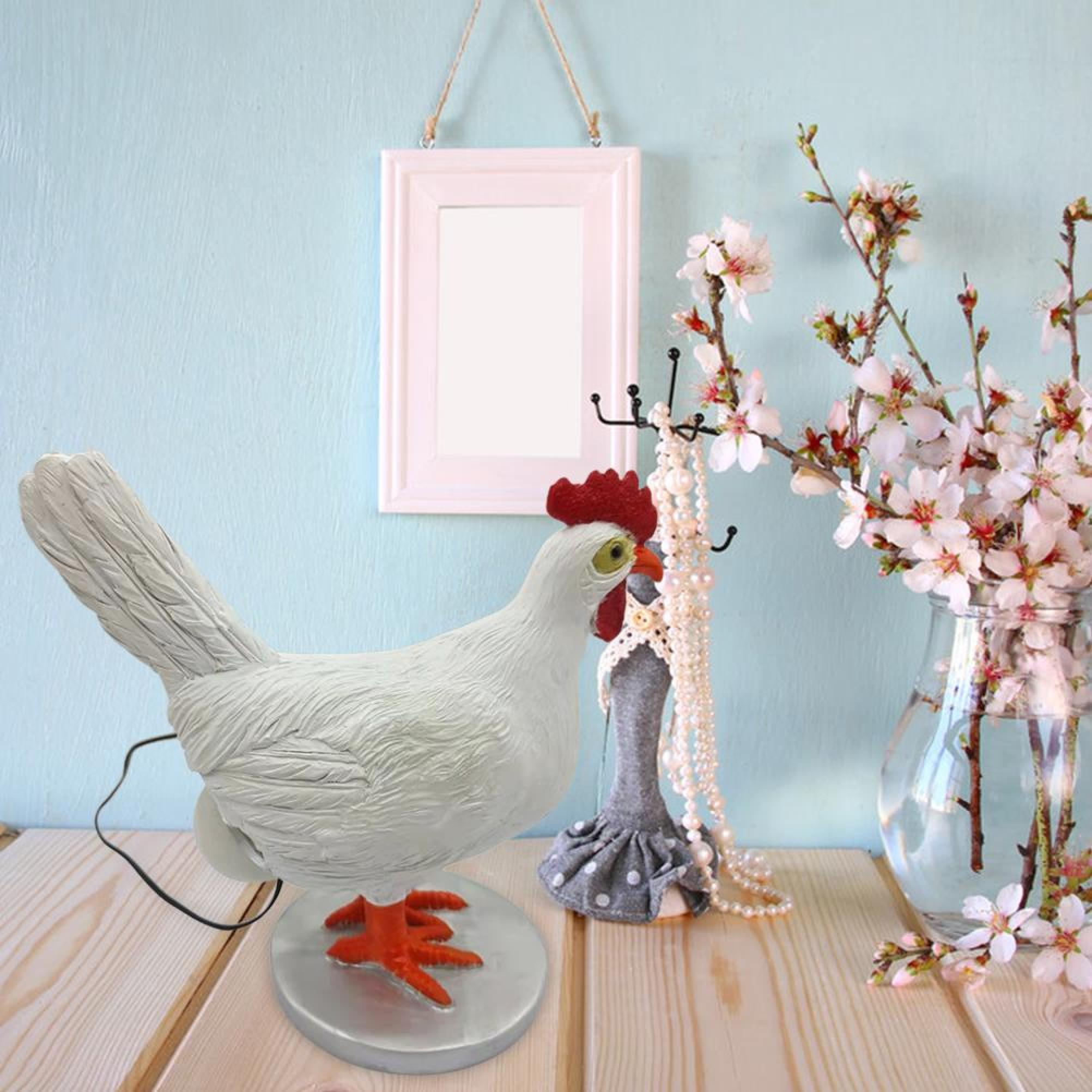 Eine weiße Hühnerfigur mit rotem Kamm auf einem Holztisch, neben einer Vase mit rosa Blüten und einem weißen Bilderrahmen.