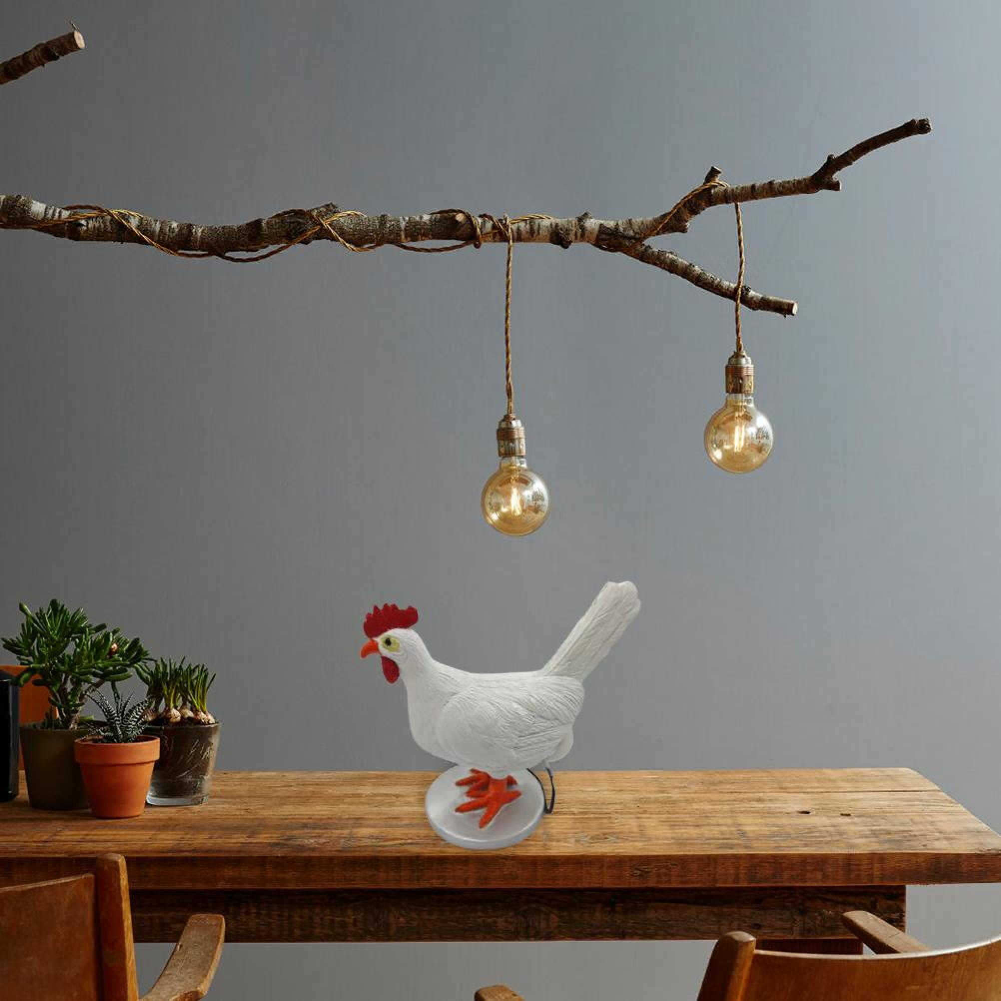 Eine weiße Hühnerfigur steht auf einem Holztisch unter hängenden Astleuchten und Topfpflanzen.