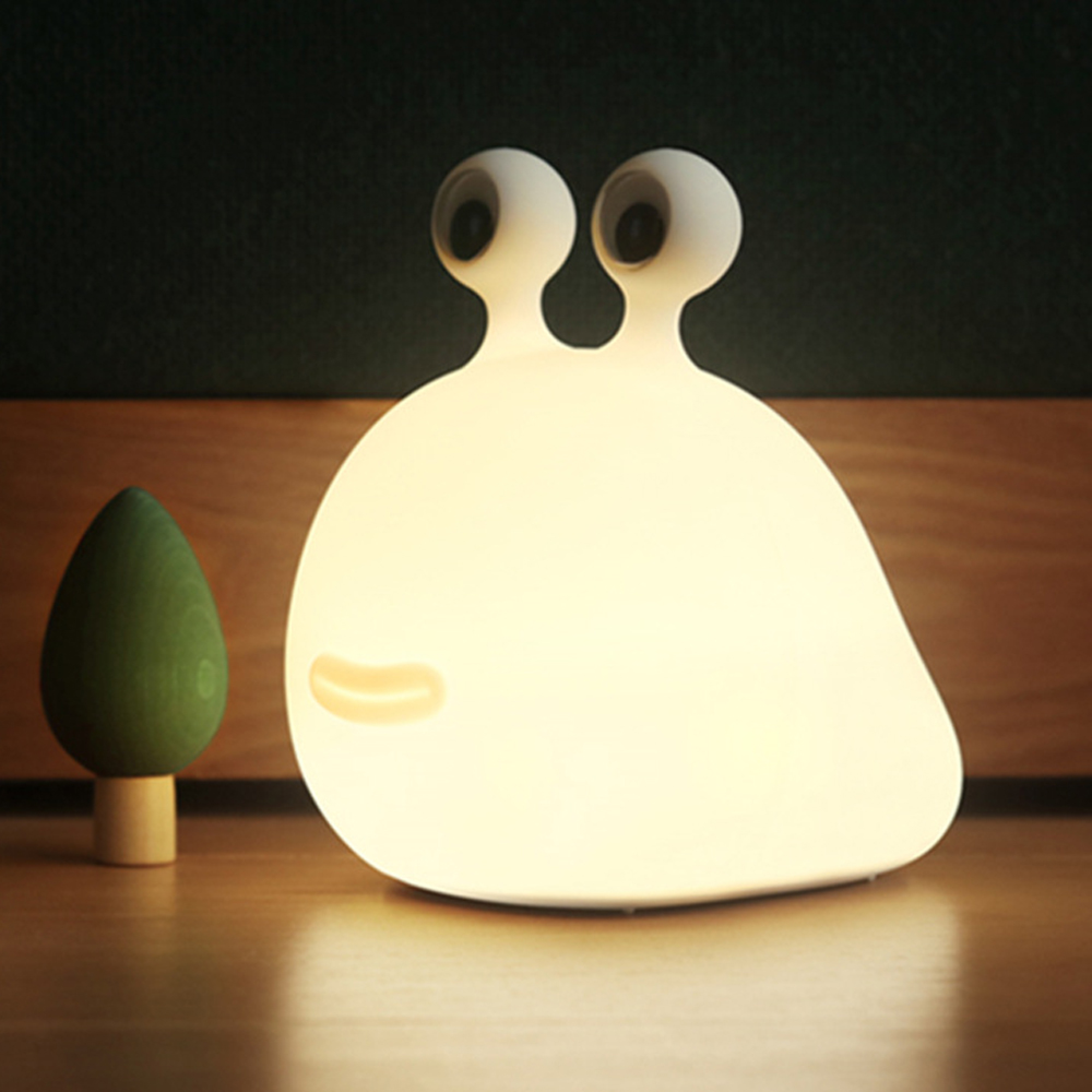 Eine weiße, leuchtende Schneckenlampe mit zwei Augen steht auf einem Tisch neben einem kleinen grünen Spielzeugbaum.