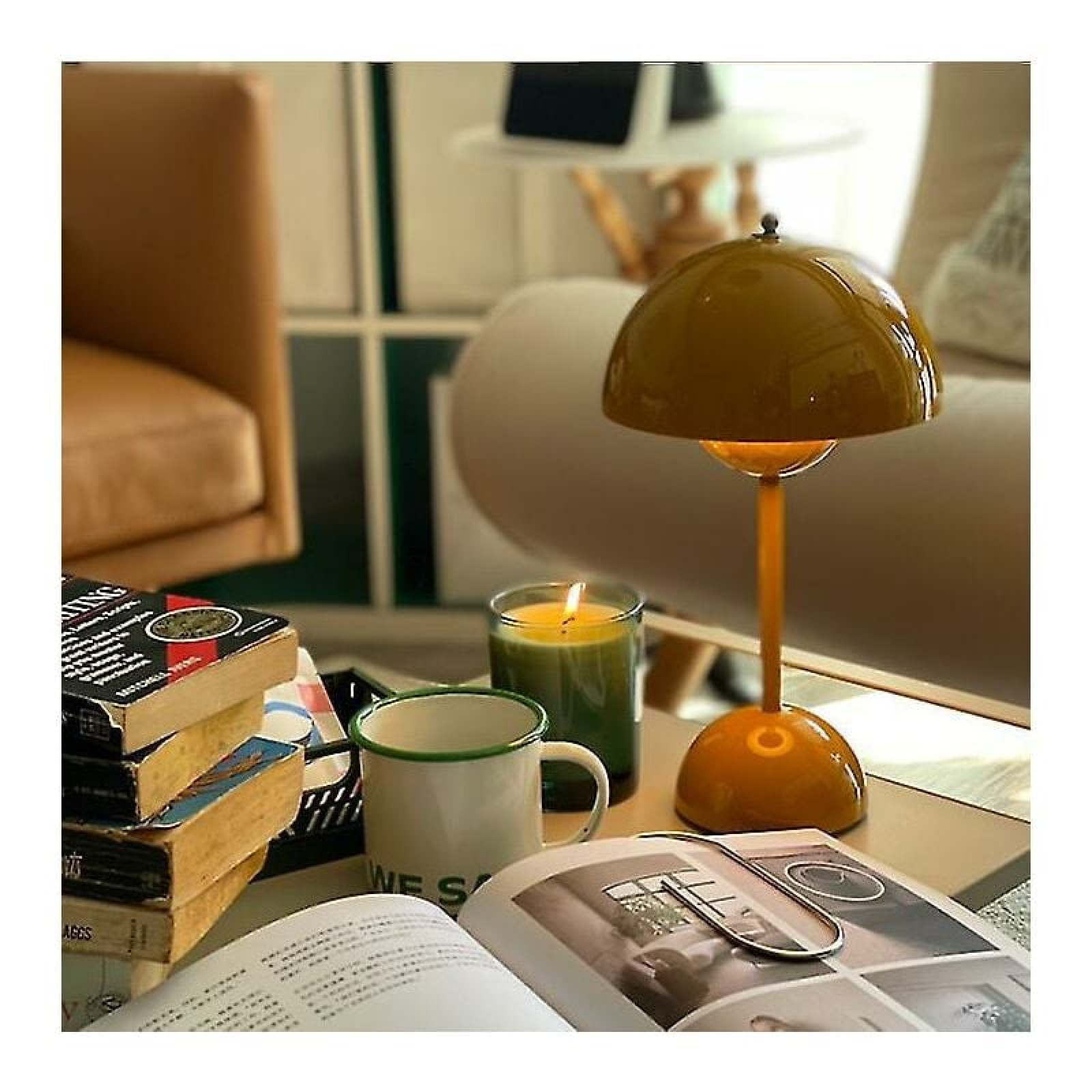 Ein Tisch mit gelber Lampe, brennender Kerze, weißem Becher, gestapelten Büchern und offenem Magazin in einem hellen Raum.