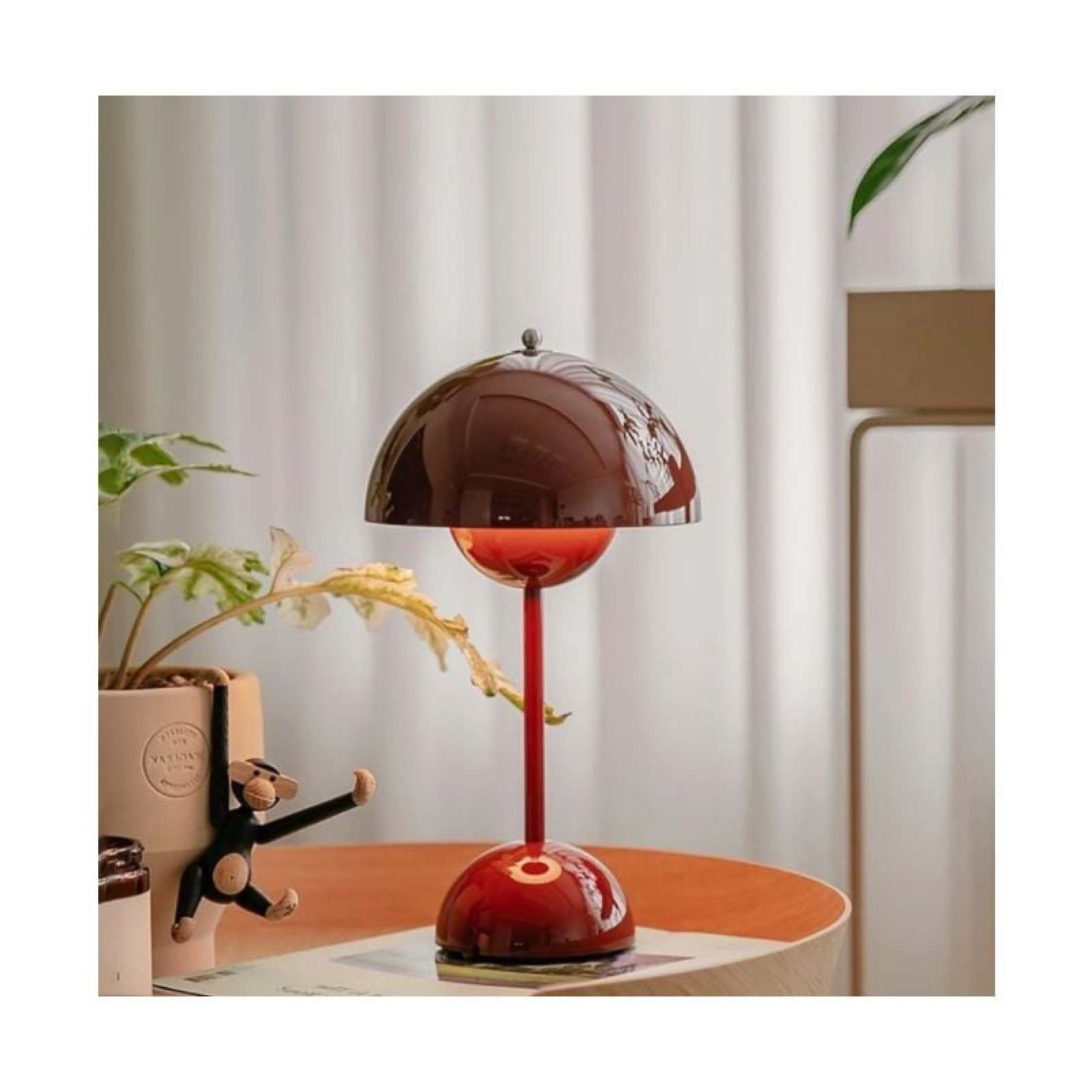 Eine glänzende dunkelrote Tischlampe mit kuppelförmigem Schirm steht auf einem Holztisch, neben Pflanze und Affenfigur.