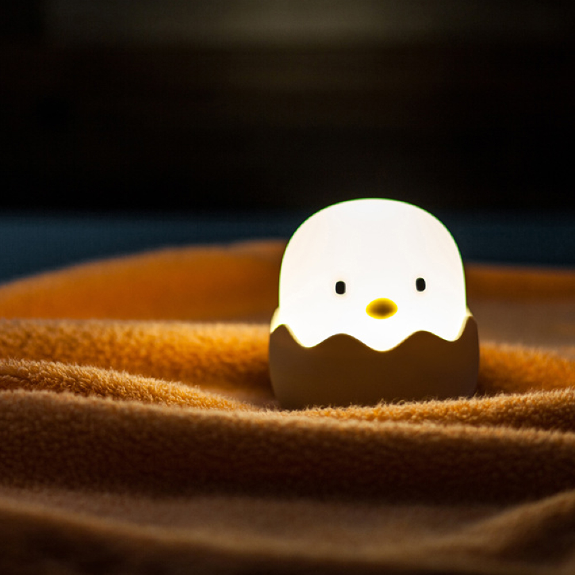 Eine kleine, leuchtende Ei-Lampe mit Gesicht sitzt auf einer weichen orangen Decke vor dunklem Hintergrund.