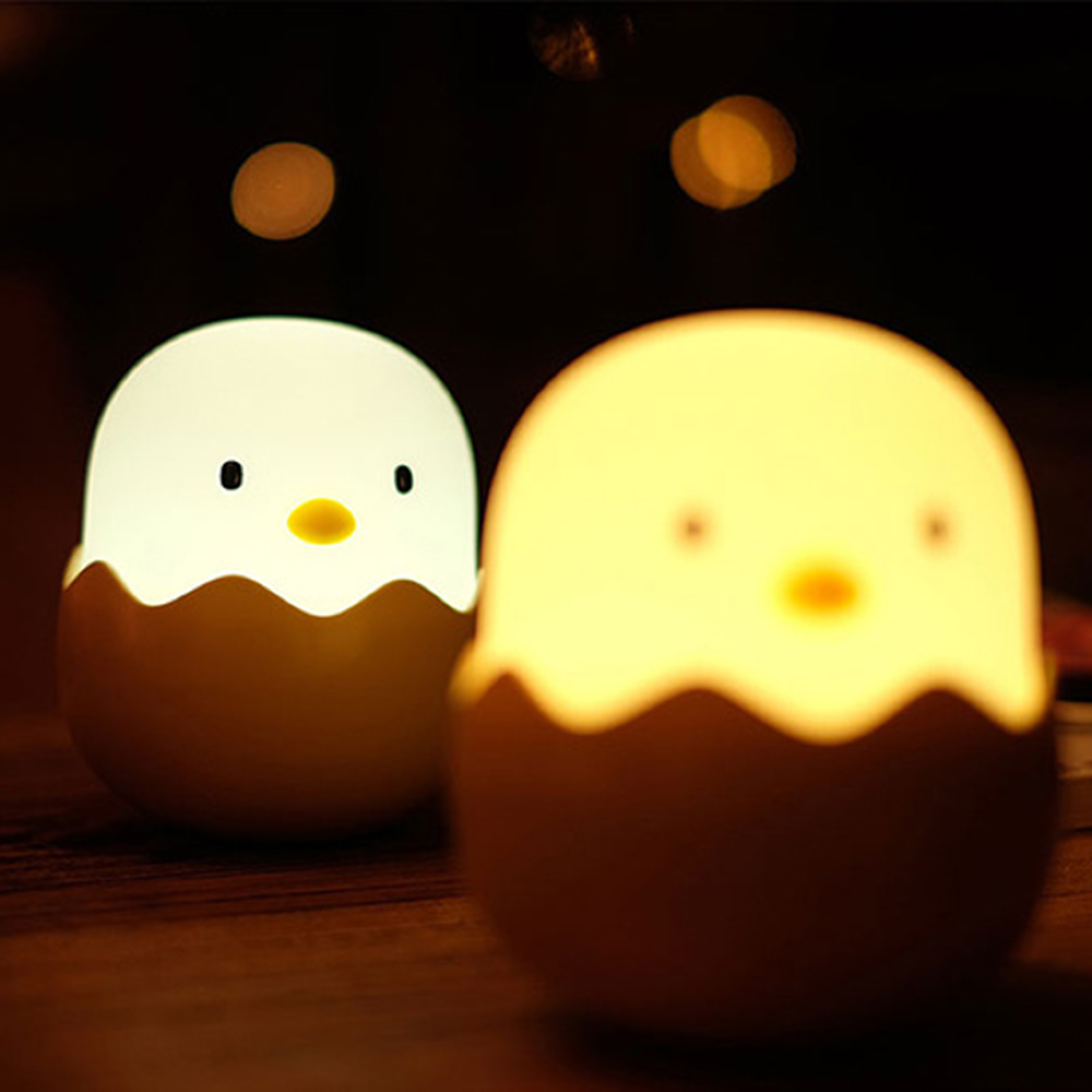 Zwei süße, leuchtende Eierlampen mit Küken-Gesichtern stehen auf einem dunklen Holztisch mit verschwommenem Hintergrund.