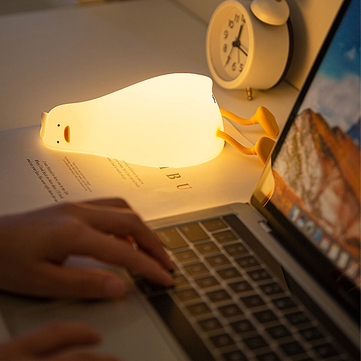 Eine Hand tippt auf einem Laptop neben einer Entenlampe und einer Uhr.