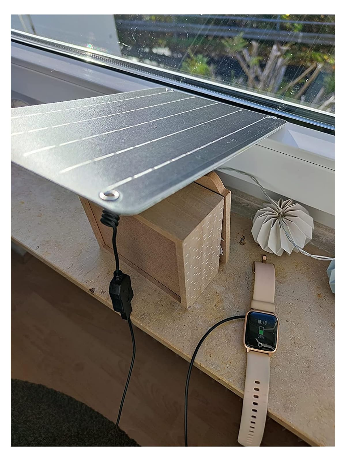 Ein kleines Solarpanel auf einer Holzkiste lädt eine Smartwatch auf einer Fensterbank.