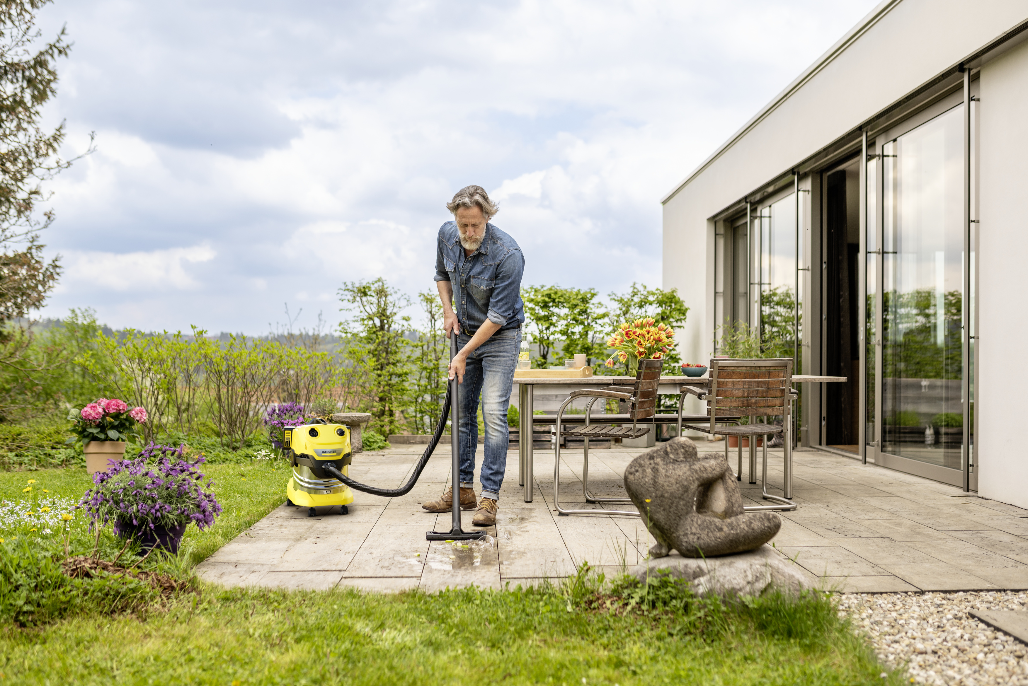 Un uomo in jeans pulisce un patio in pietra con un aspiratore per liquidi giallo fuori da una casa moderna.