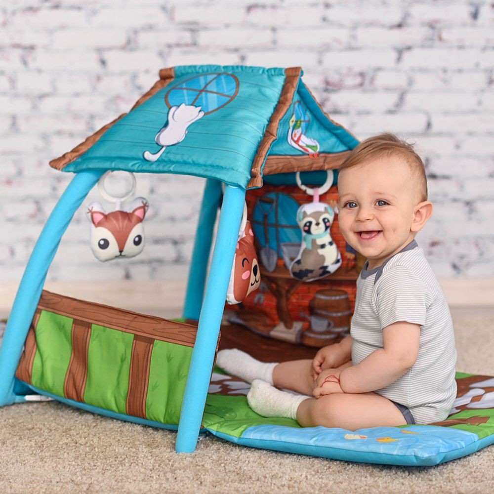 Ein lächelndes Baby sitzt auf einer Spielmatte neben einem blauen Stoffzelt mit hängenden Kuscheltieren.