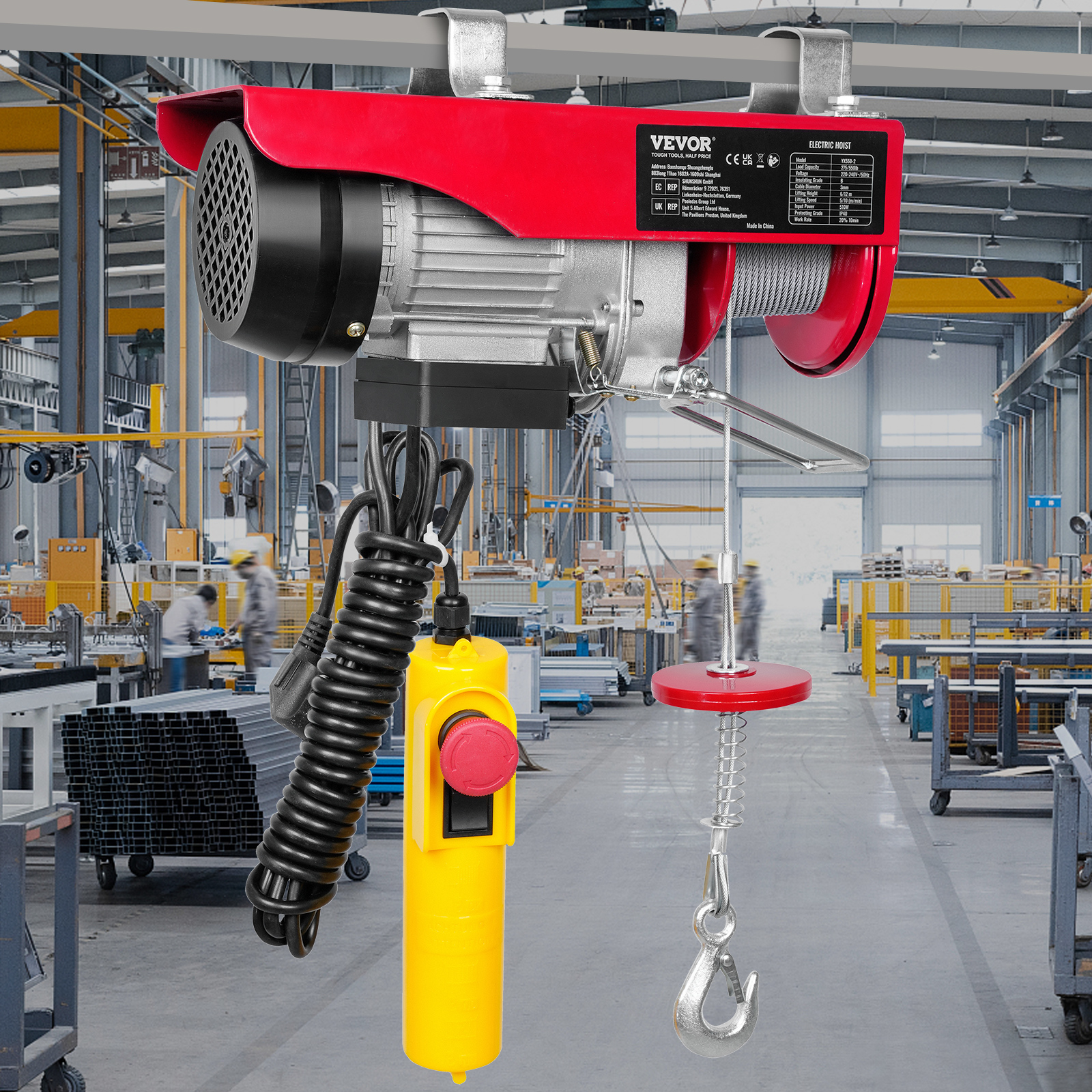 Un polipasto eléctrico rojo y plateado cuelga en una gran fábrica industrial con estructuras metálicas.