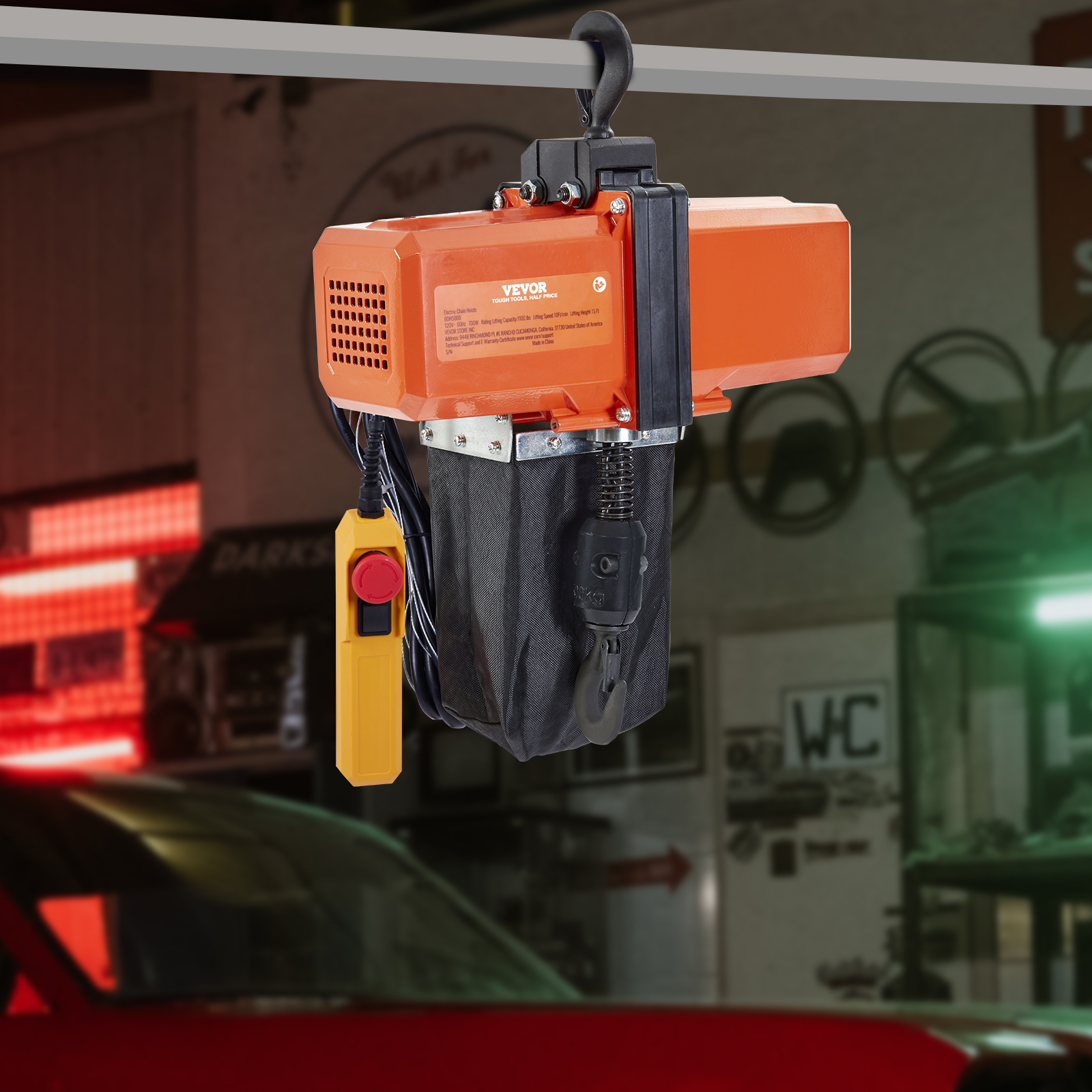 Un polipasto eléctrico naranja cuelga de una viga con un control amarillo en un taller.