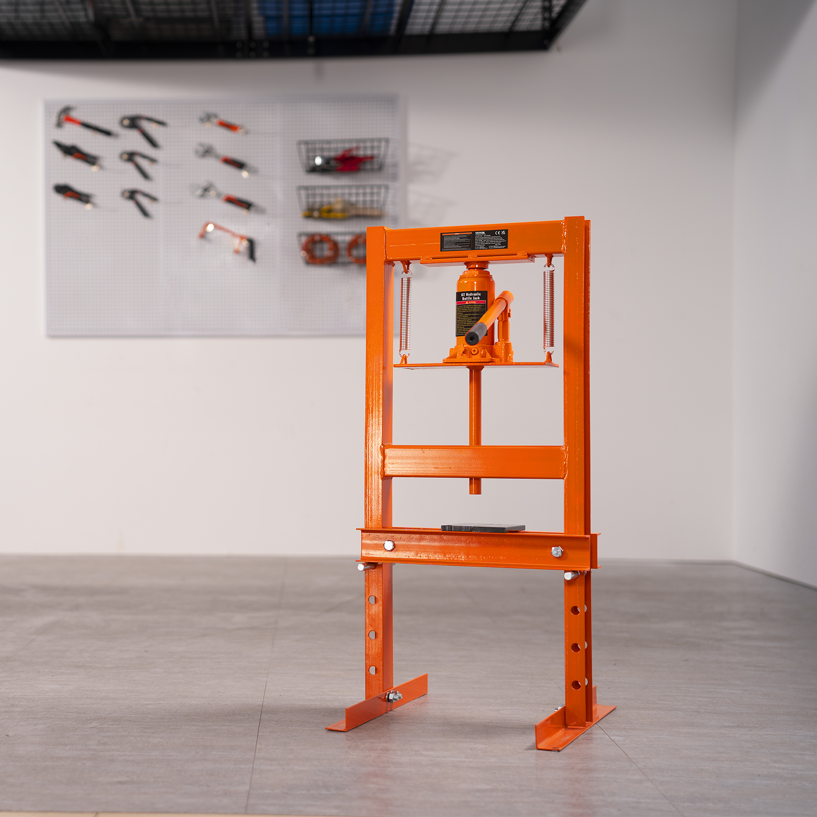 Una prensa de taller de color naranja brillante sobre un suelo gris, con un panel de herramientas al fondo.