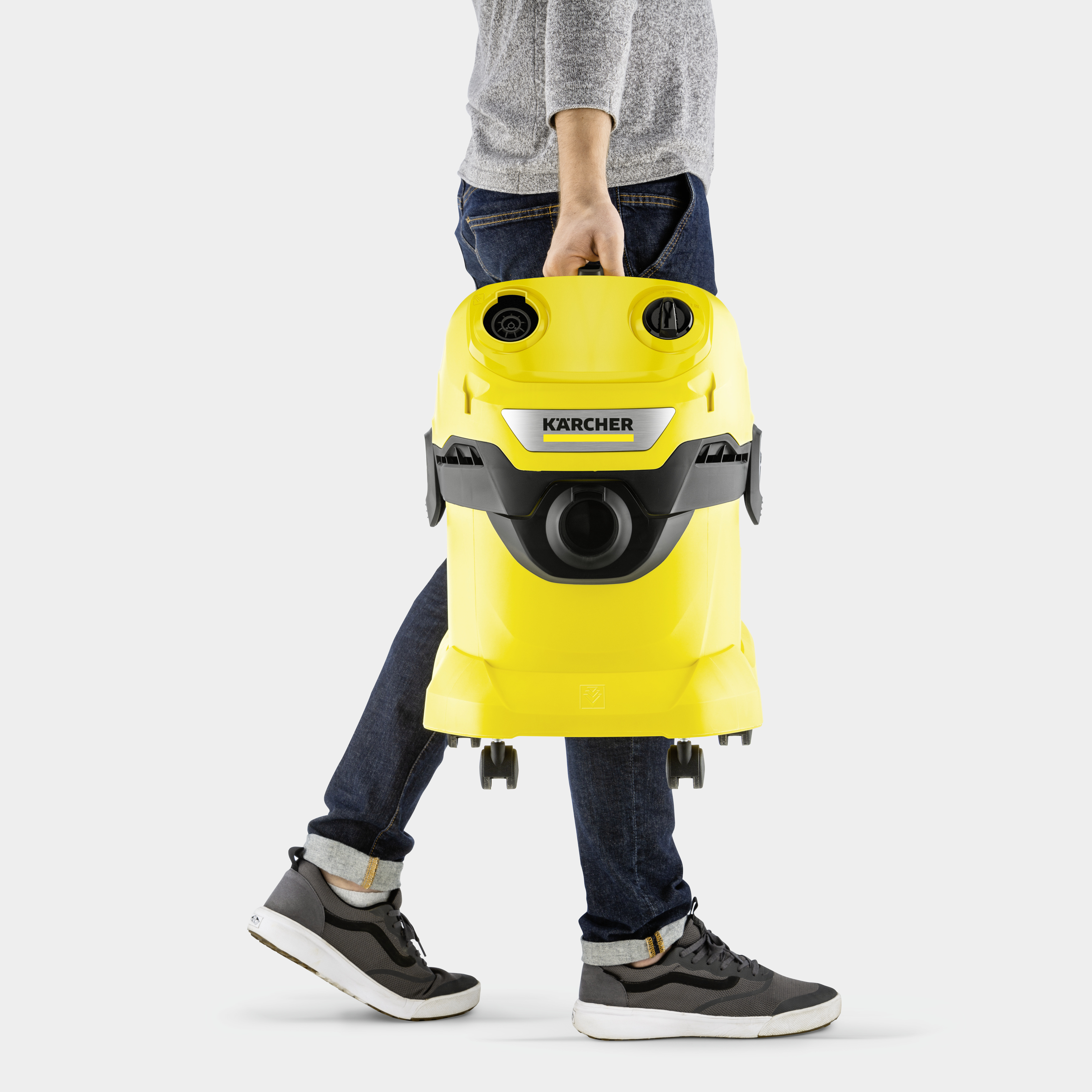 Una persona con jeans e maglione grigio trasporta un aspirapolvere Kärcher giallo.