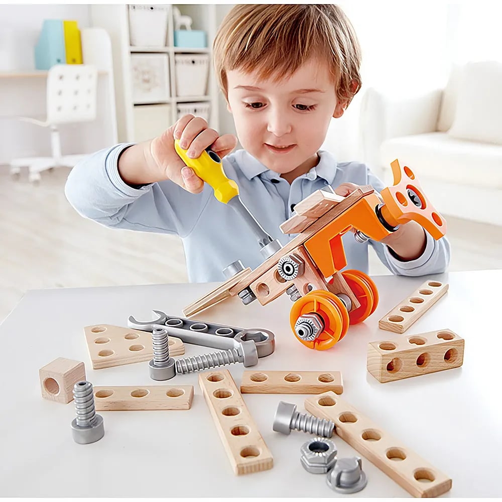 Kleiner Junge sitzt am Tisch und baut mit Holzteilen und Werkzeug ein Spielzeugfahrzeug zusammen.